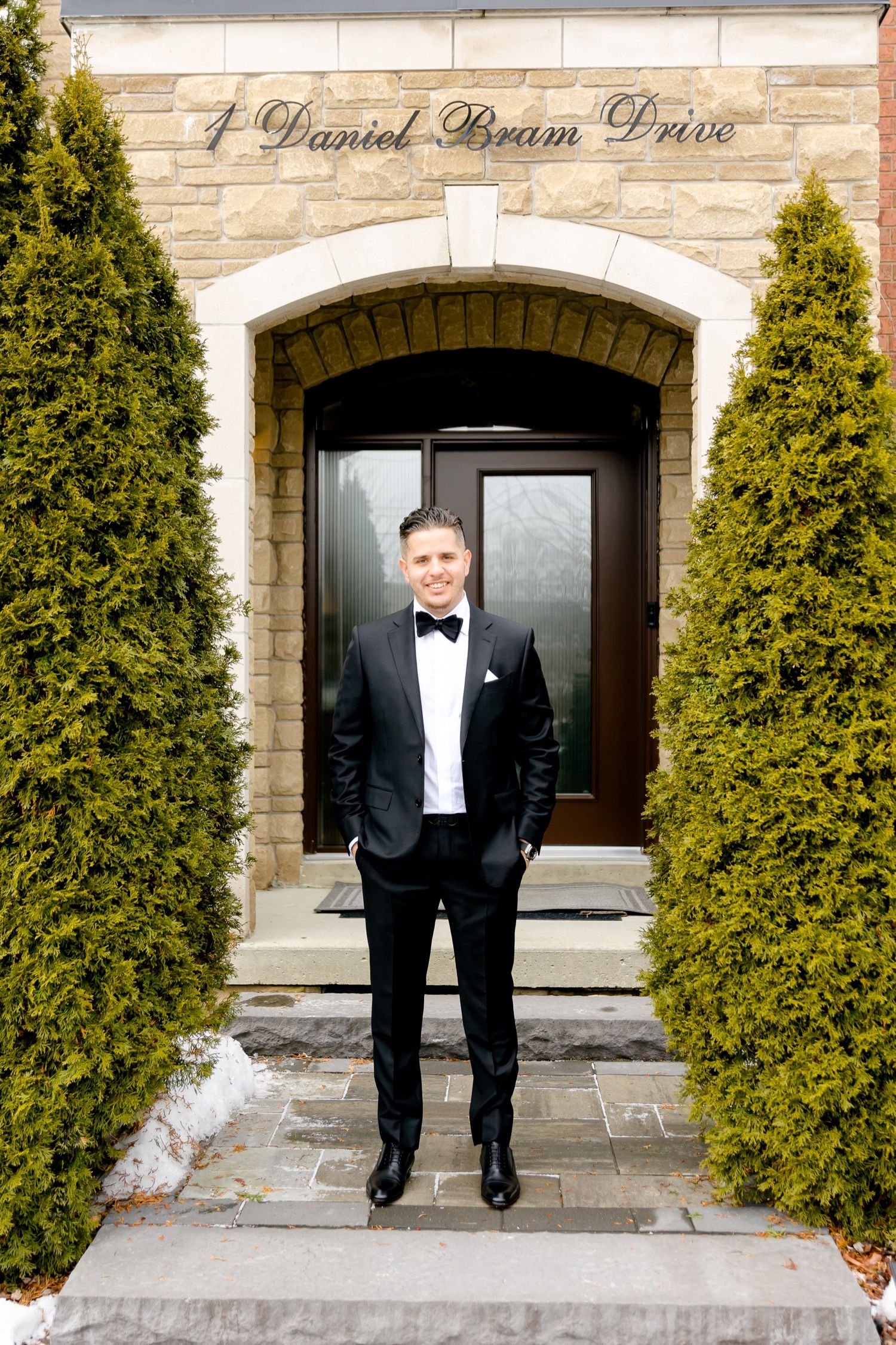 Groom in tuxedo stands between cypress trees at formal entrance.