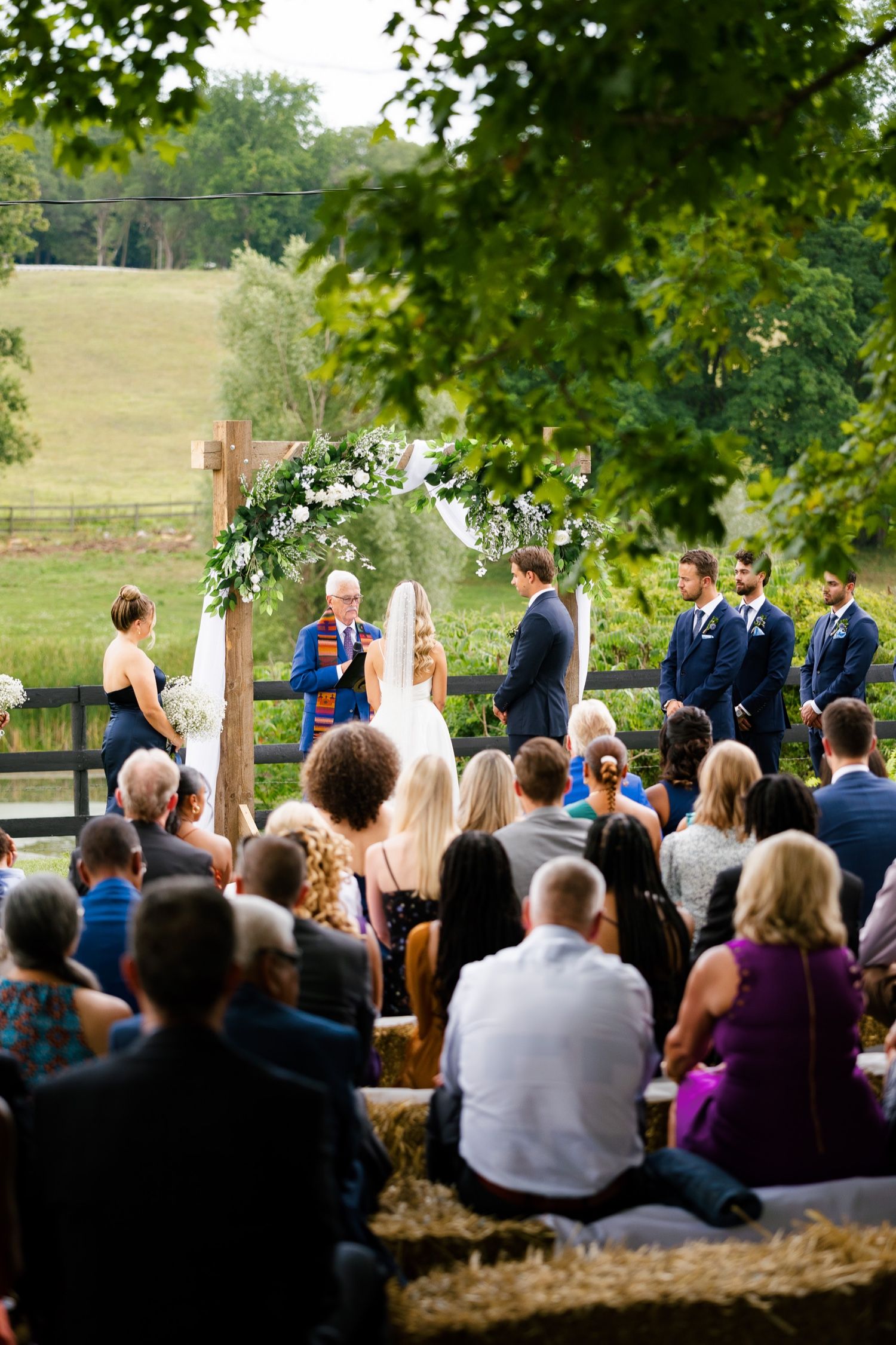 Guests seated on hay bales watch an outdoor wedding ceremony under a floral arch.