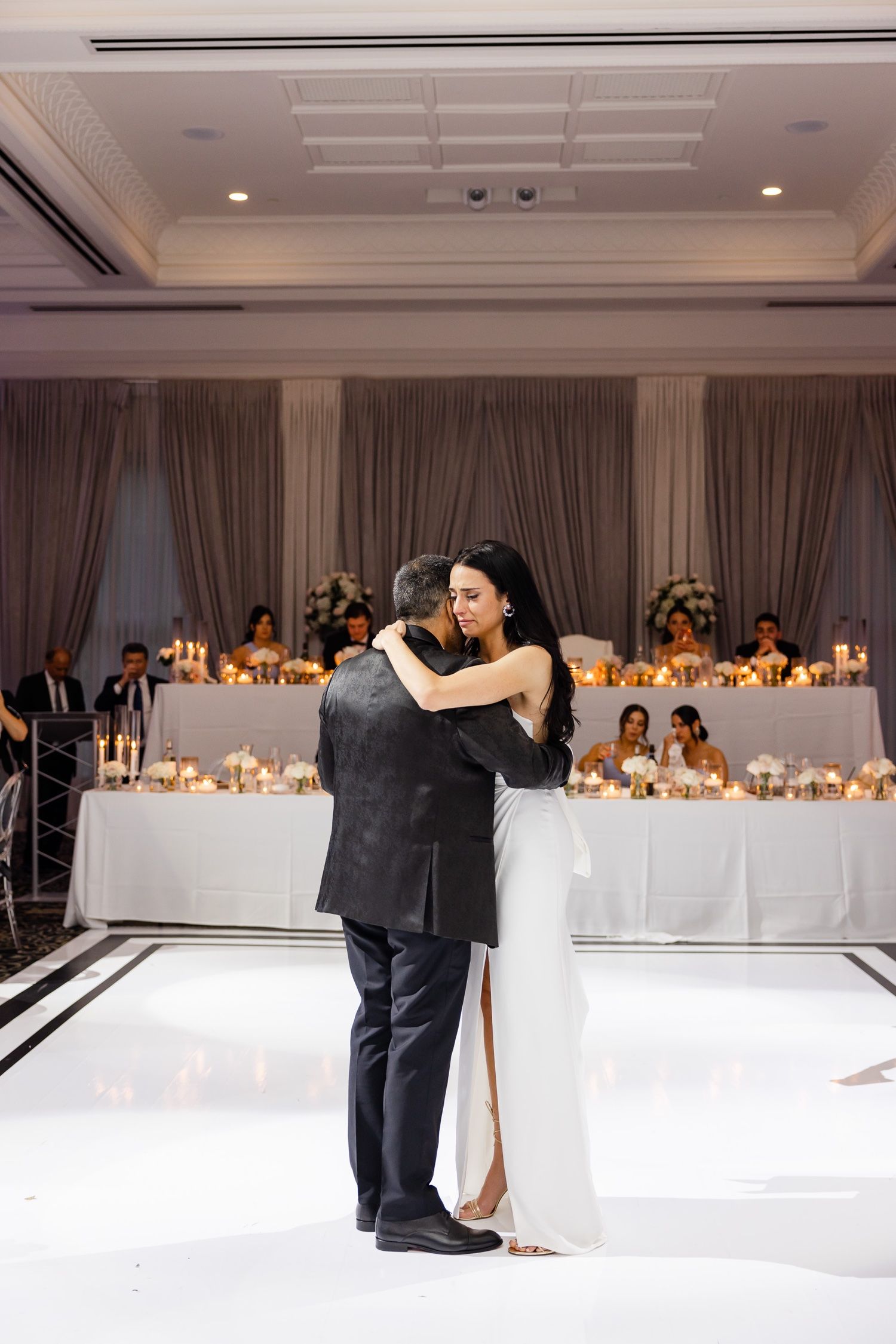 Couple embraces during romantic first dance on white dance floor with candlelit reception tables behind them.
