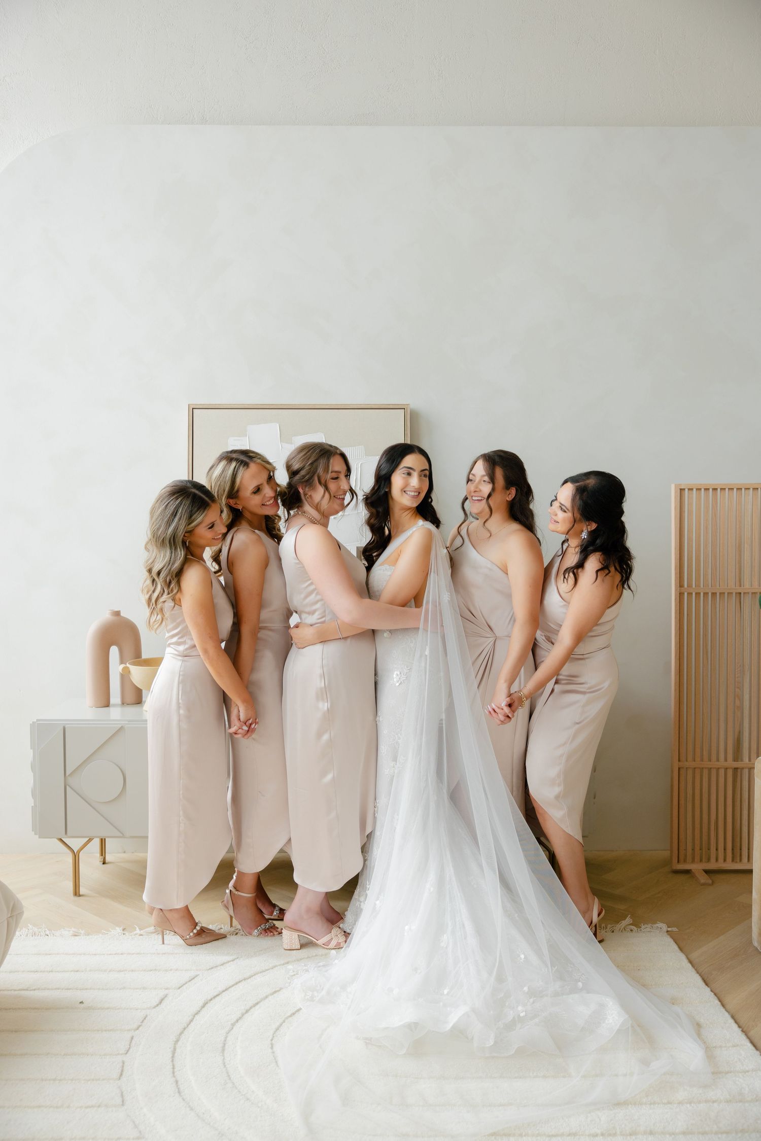 Bridal party in neutral dresses gathered around bride in elegant white gown.