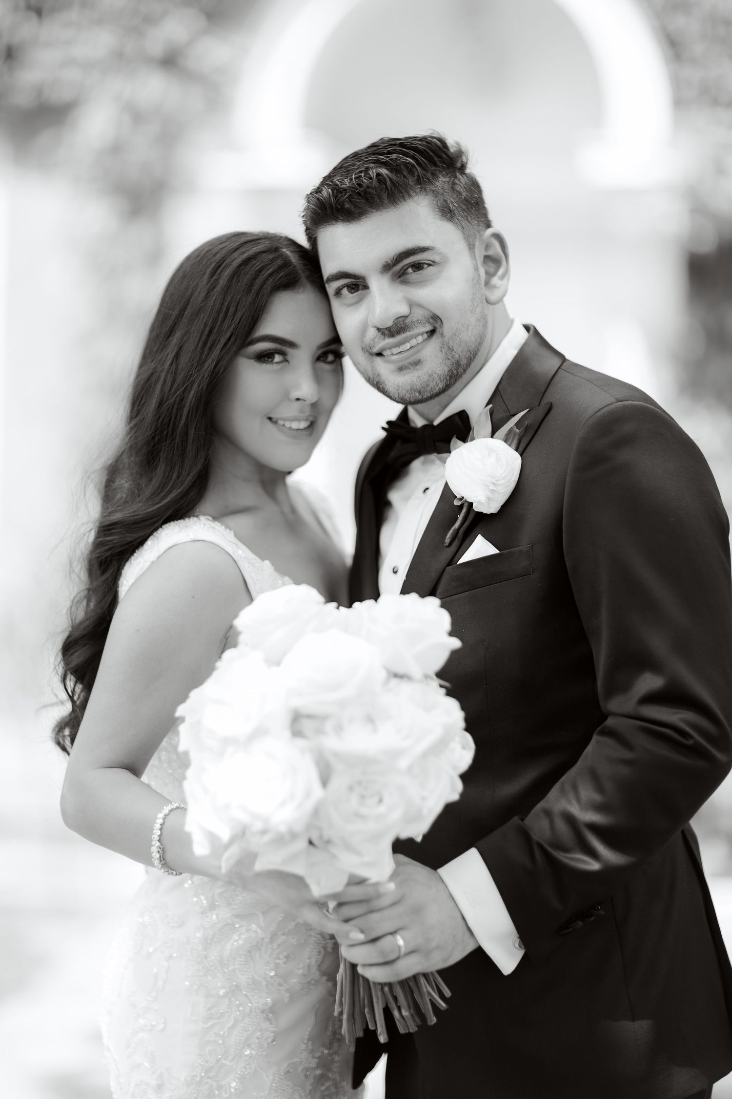 Classic black and white portrait of newlyweds with white rose bouquet under garden arch.