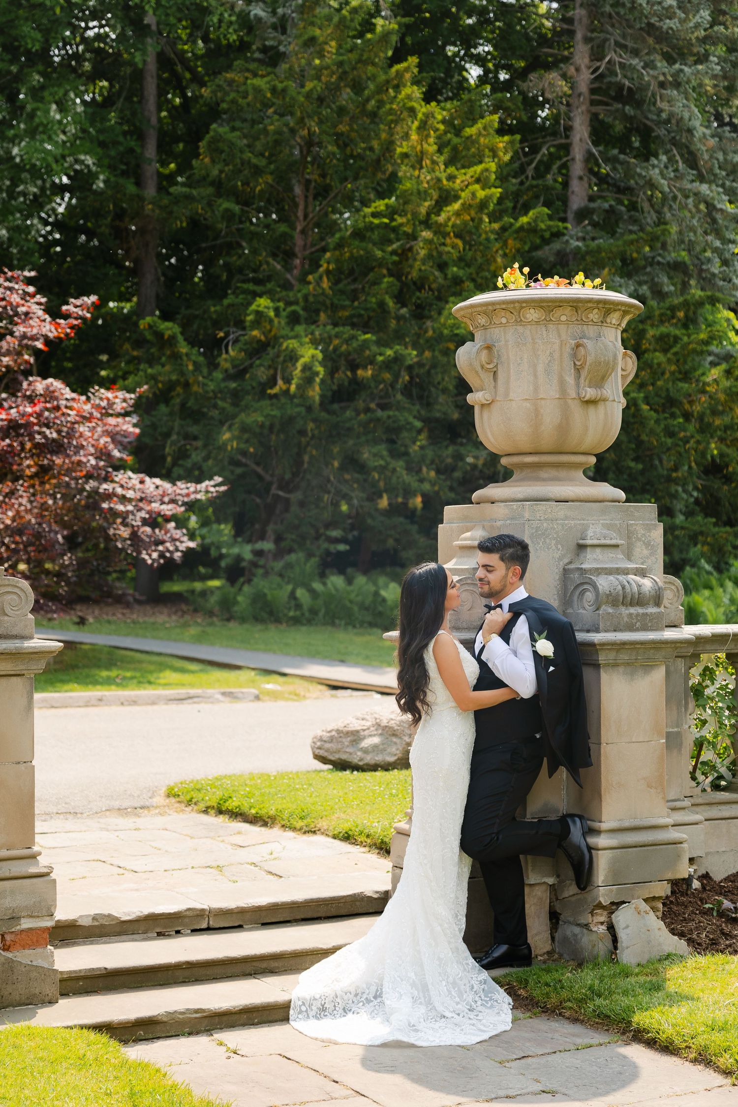 Elegant wedding couple poses by ornate garden urn on manicured grounds.