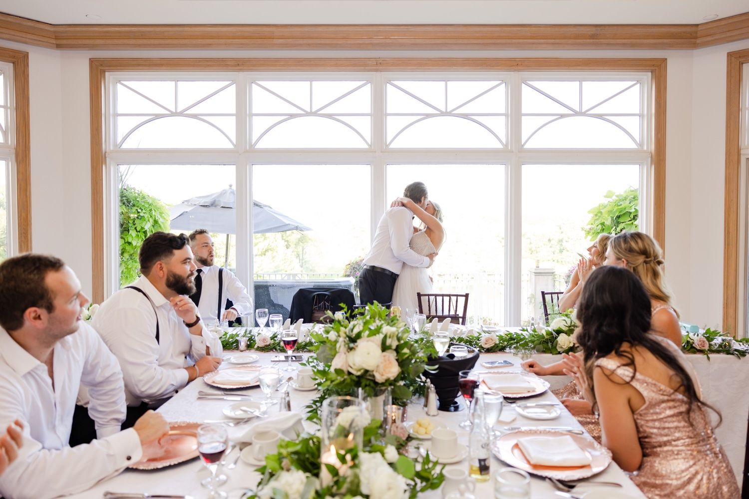 Wedding guests enjoy an elegant reception dinner with floral centerpieces in a bright windowed venue.