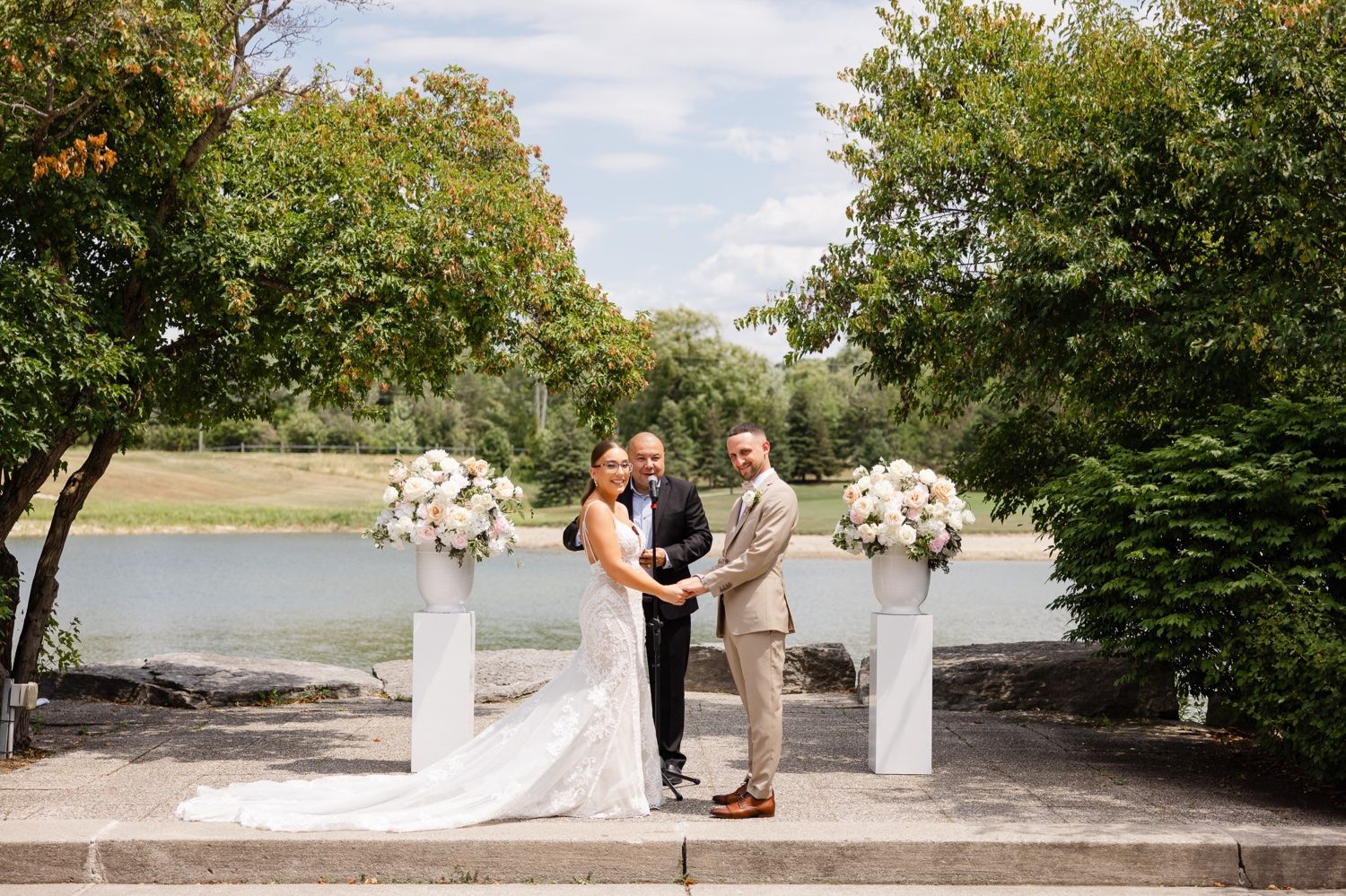 Wedding ceremony takes place on stone patio with lakeside view and elegant white floral decor.