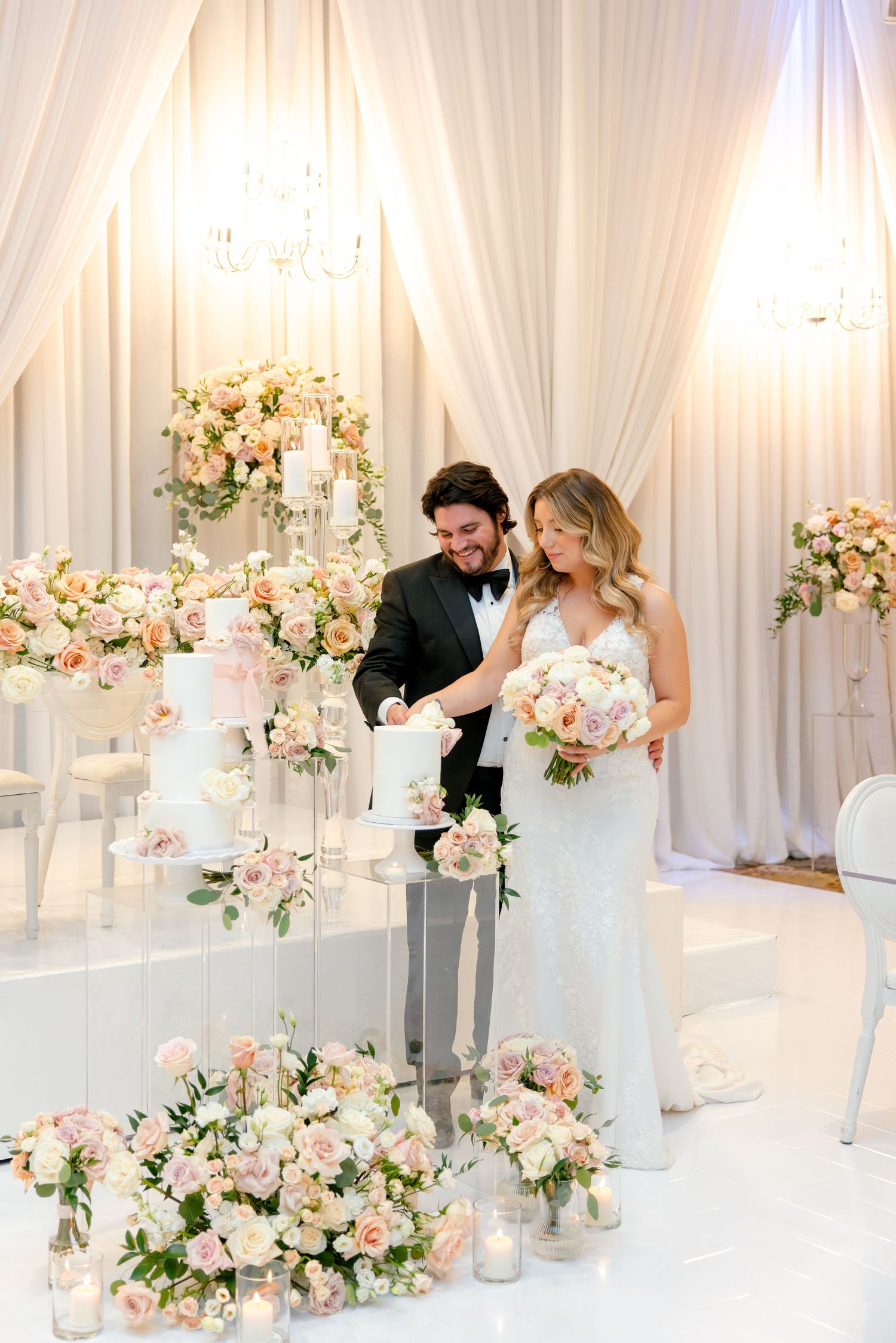 Elegant wedding cake display surrounded by blush floral arrangements and white candles.