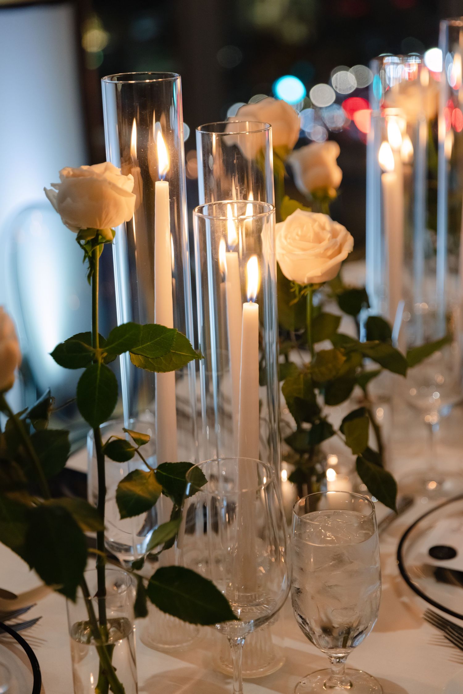 Elegant table setting with tall glass candleholders and cream roses creating a romantic ambiance.