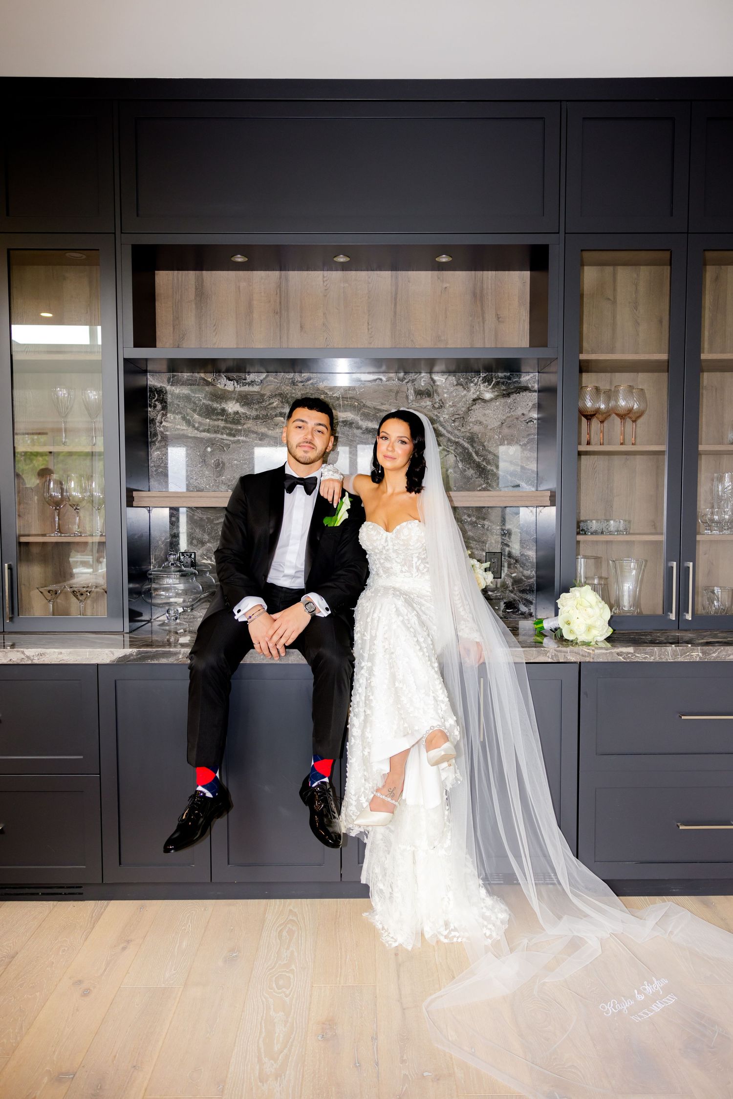 A couple poses together in a modern kitchen with dark cabinets and glass displays during their wedding photoshoot.