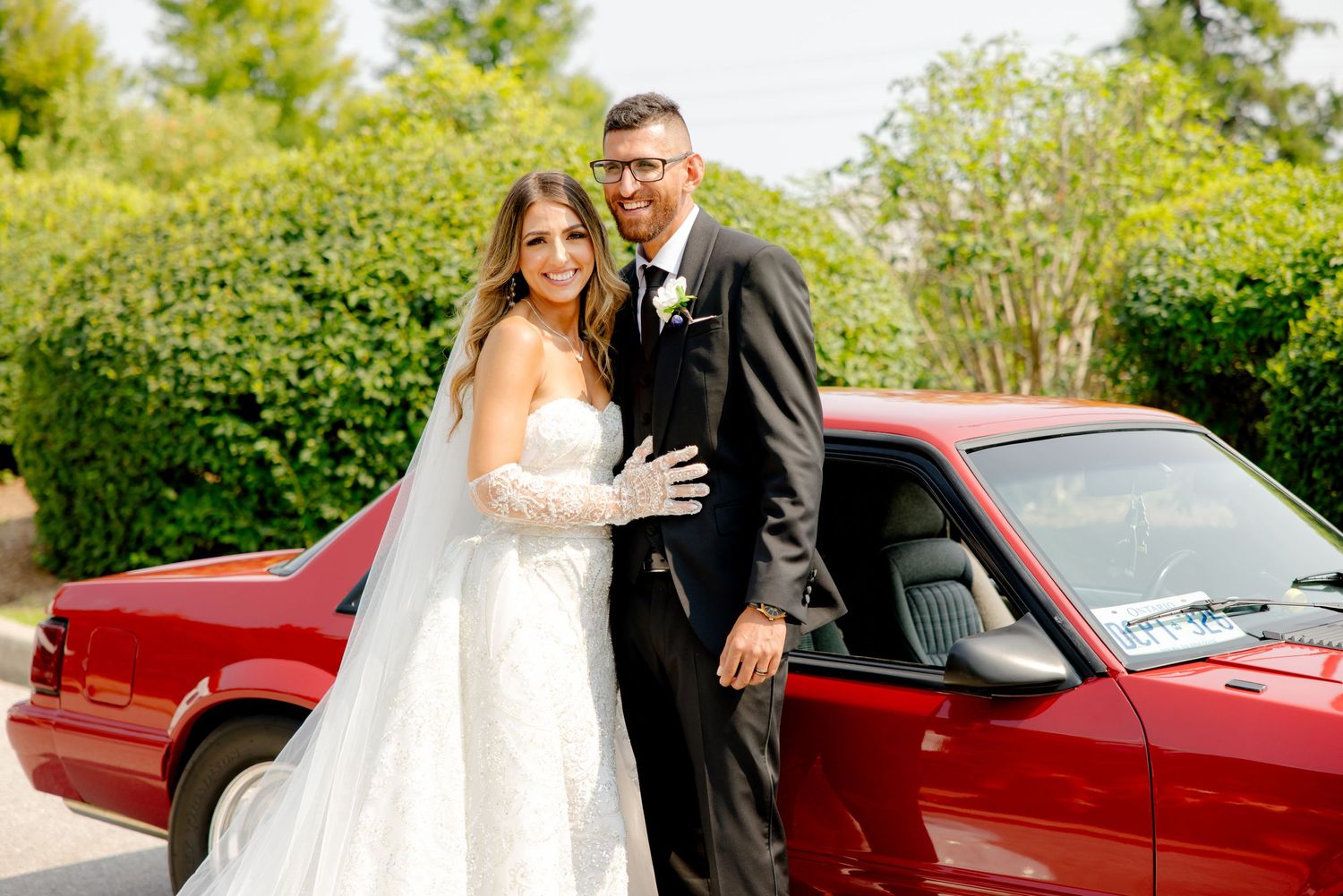 A beautiful wedding photo featuring a classic red car with a bride in a white gown and groom in a black suit.