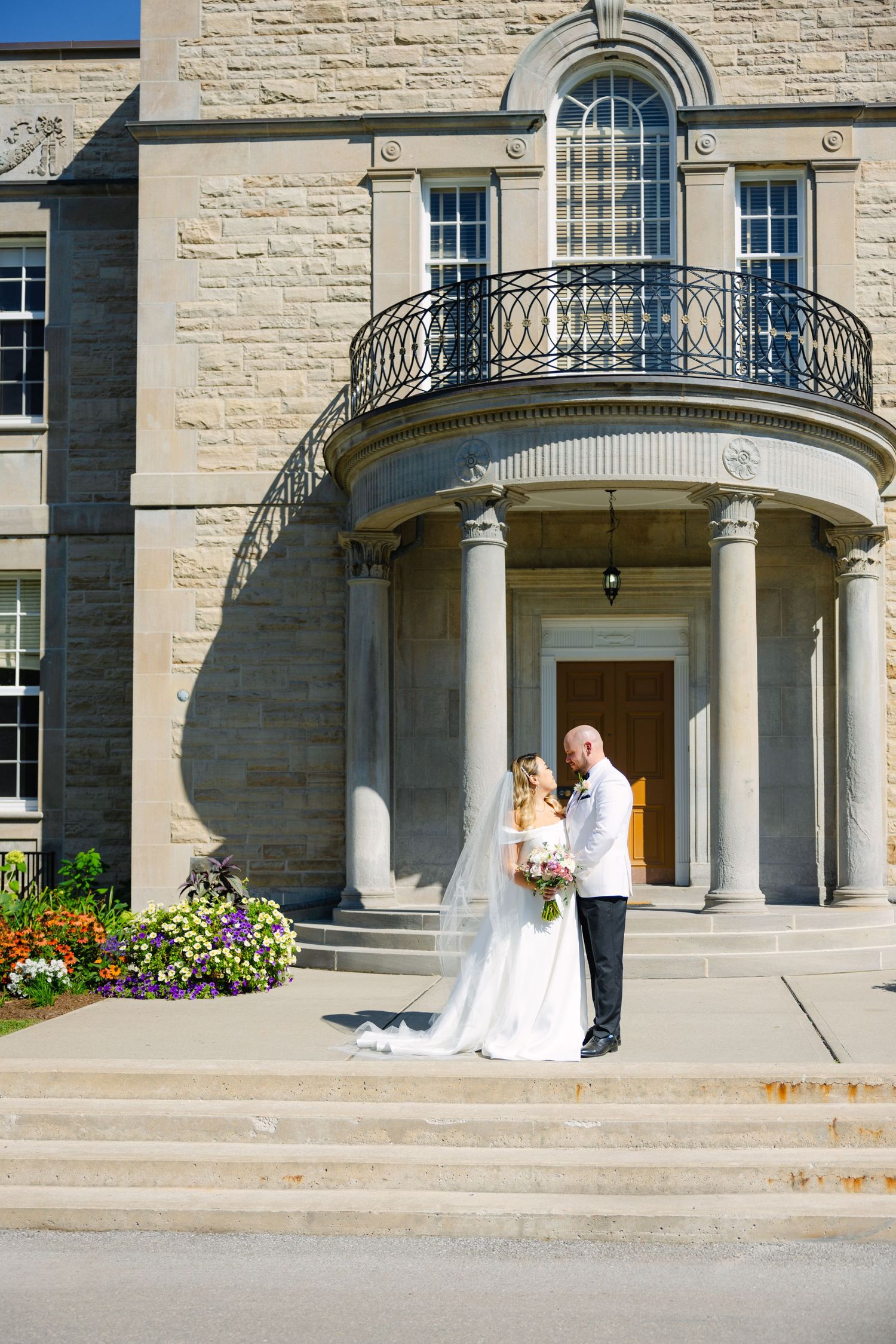 Couple in wedding attire standing on steps of an elegant stone building with curved balcony.