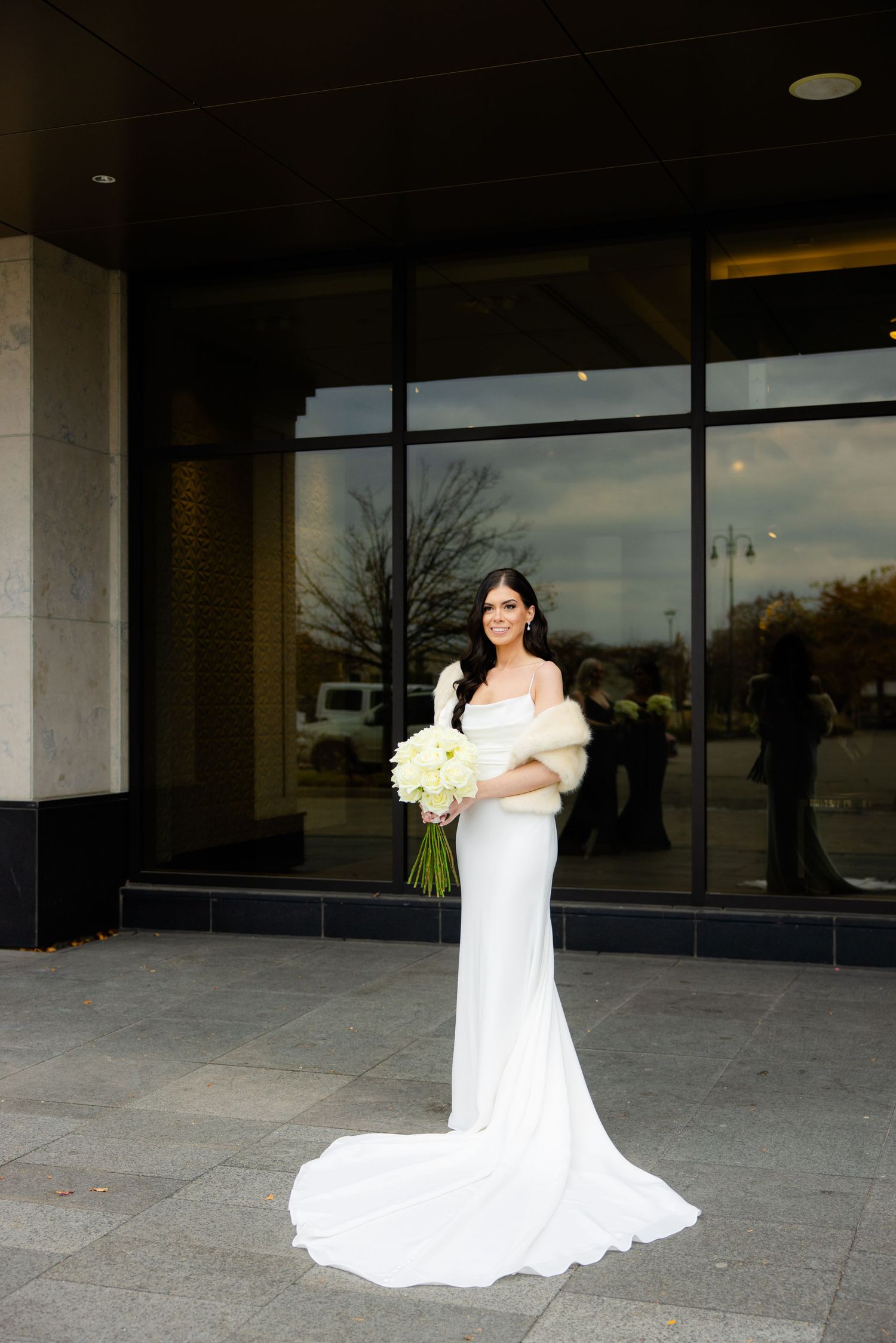 Bride in sleek white wedding dress with fur wrap holds cream bouquet against modern building backdrop.