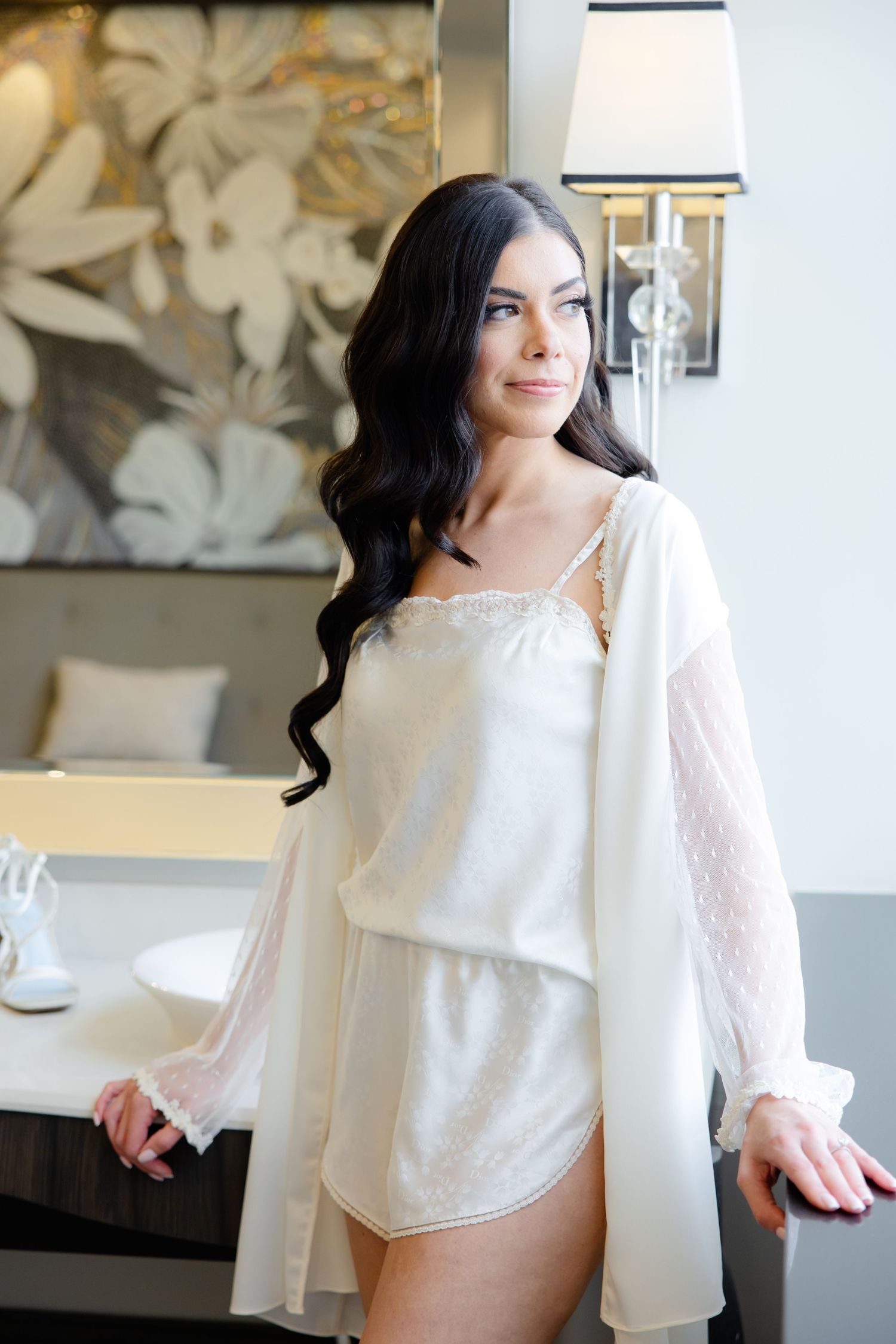 Bride in elegant white robe stands beside decorative floral wallpaper during wedding preparations.