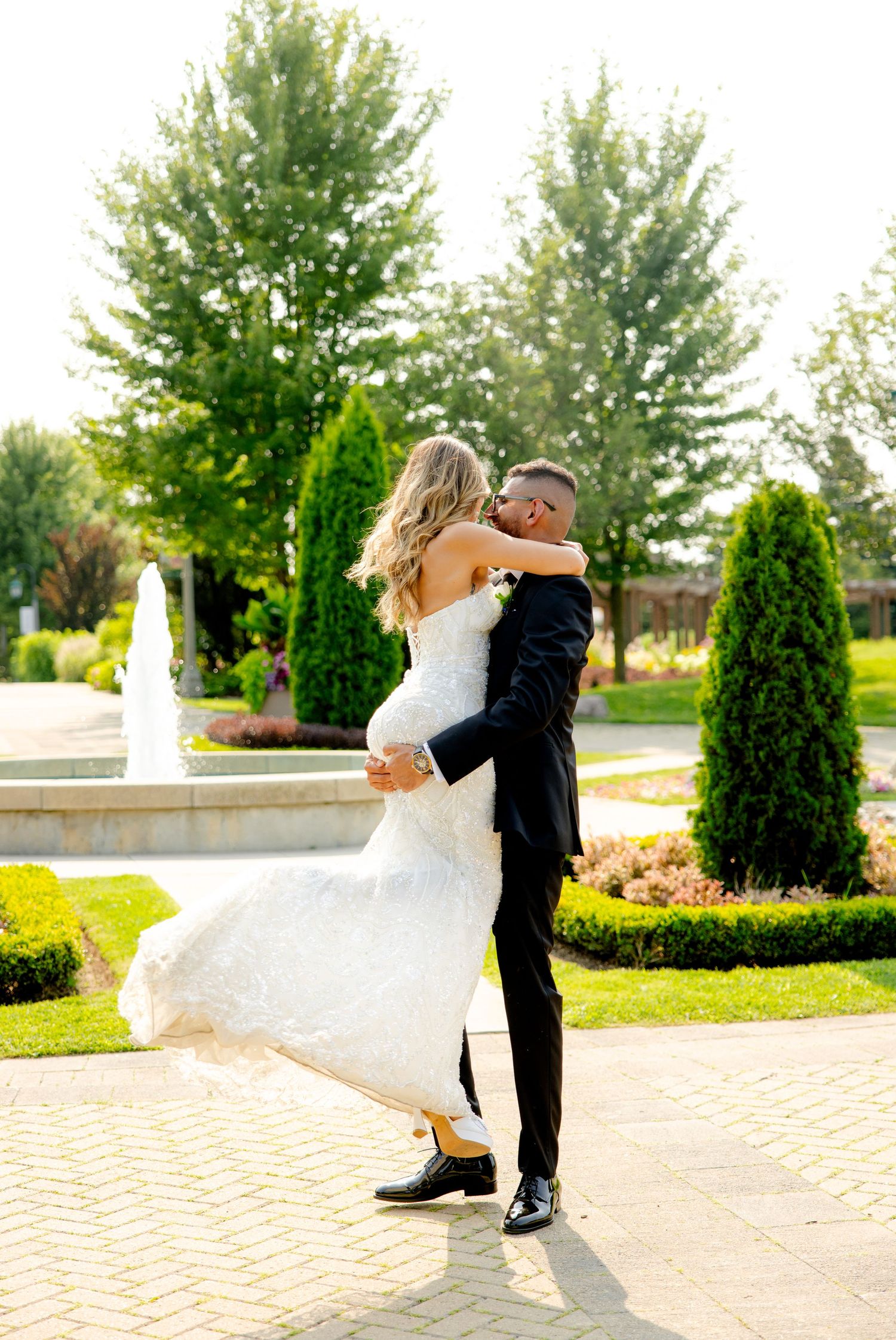 Romantic wedding photo of couple embracing in a garden setting with fountain and evergreen trees.