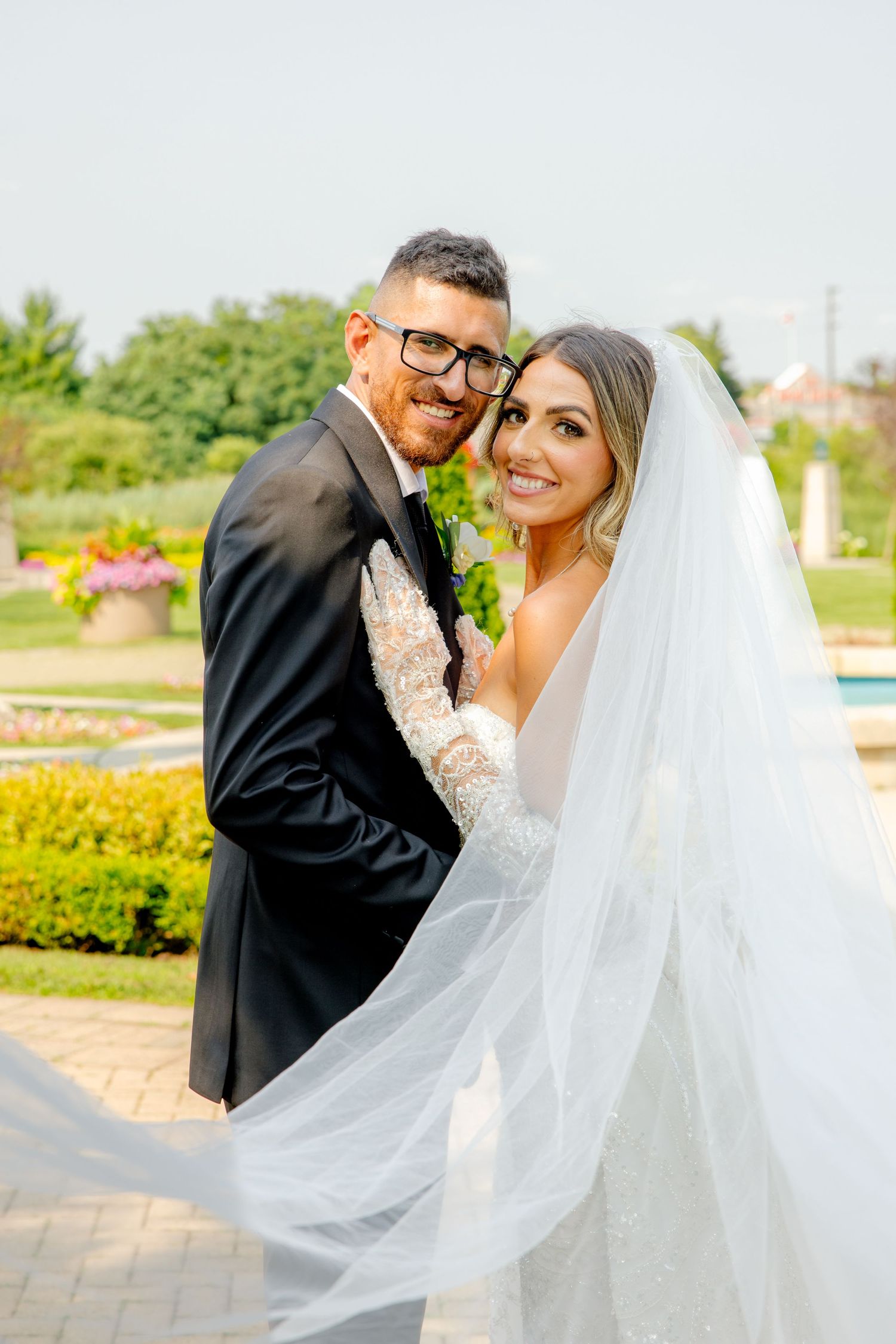 Joyful wedding portrait captured in landscaped garden with flowing veil and evergreen backdrop.