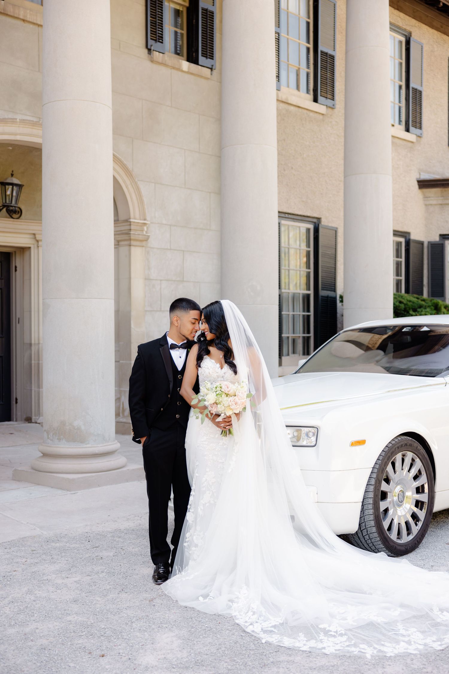 Elegant wedding photo with couple embracing near white car and stone columns.