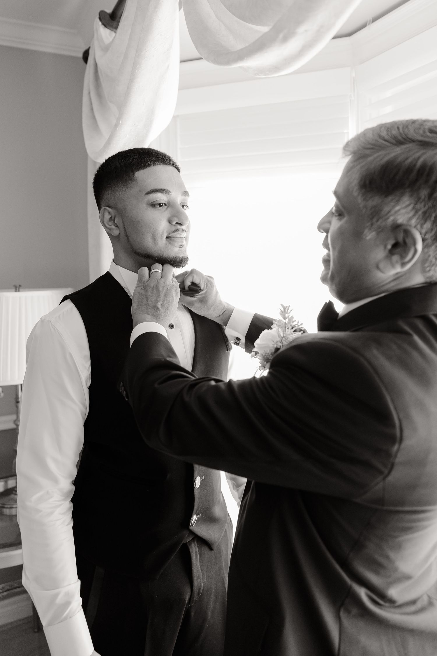 Two people in formal attire make final adjustments to clothing near a window with draped curtains.