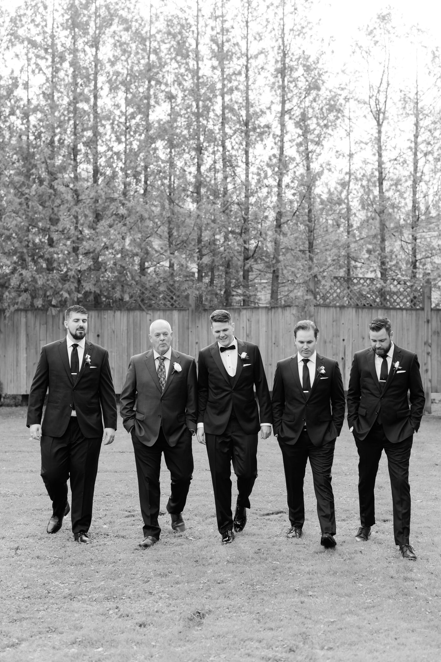 Black and white photo of five people walking together in formal attire against a wooded backdrop.