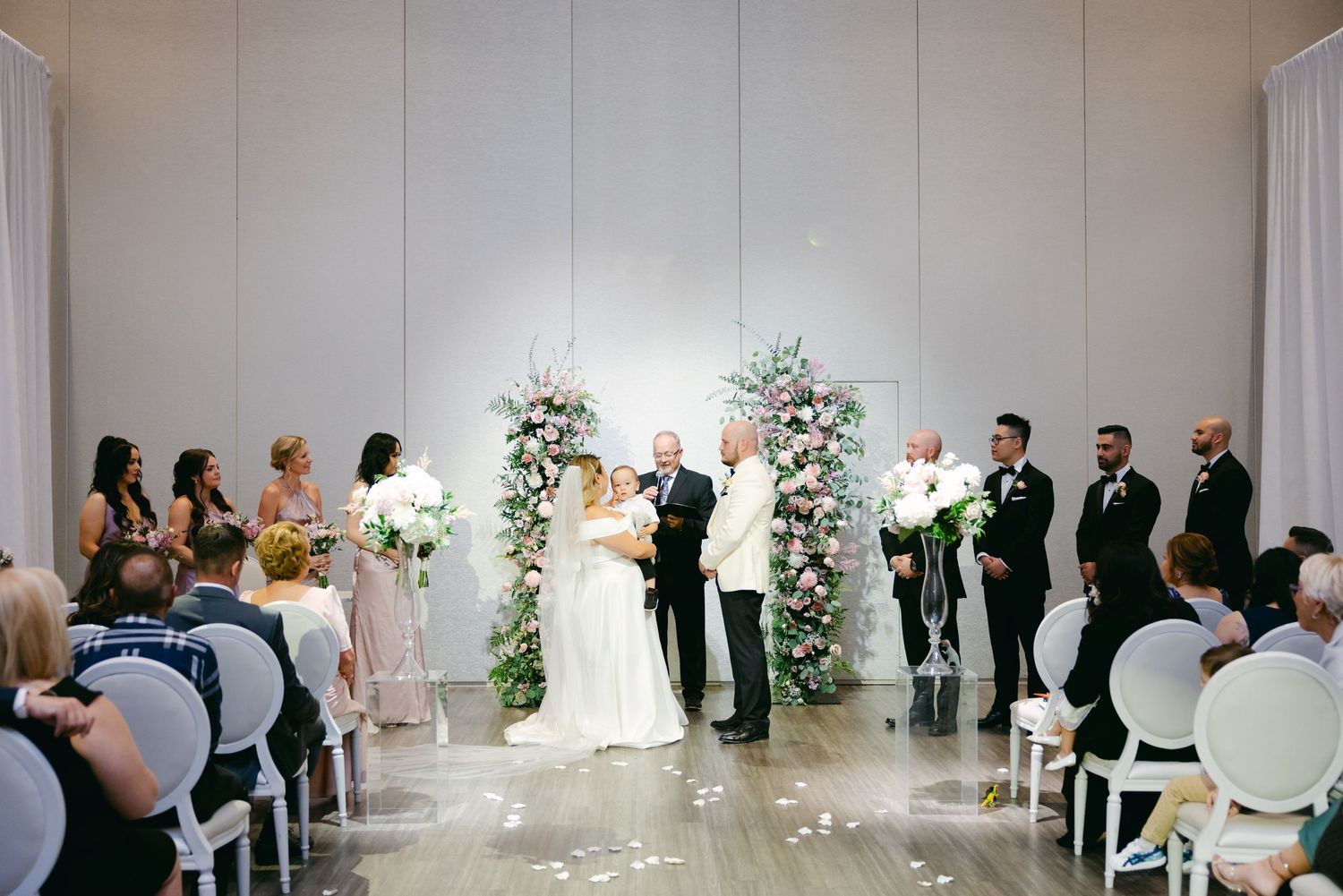 Wedding ceremony in elegant white room with floral arch and guest seating.