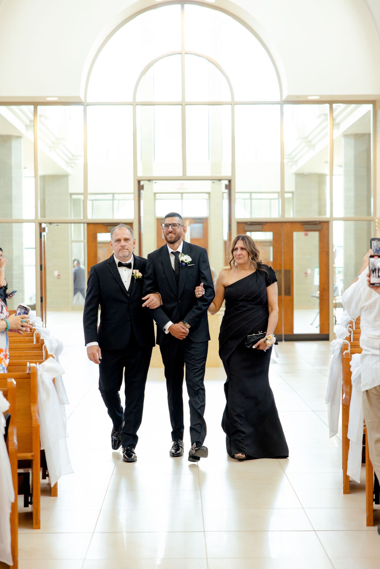 Three people walk down a church aisle with arched windows and wooden pews.