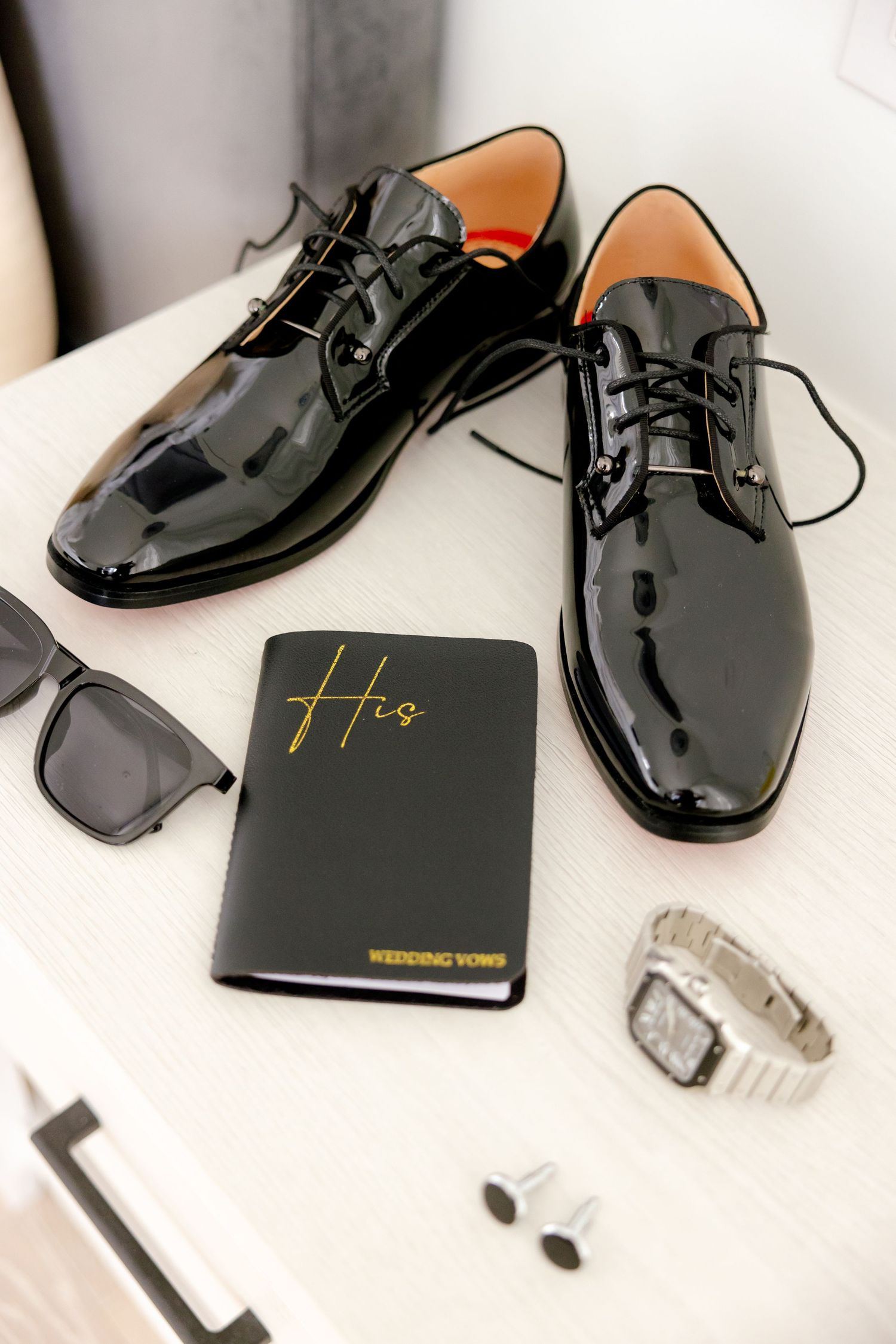 Patent leather dress shoes displayed with sunglasses, watch and black passport holder.