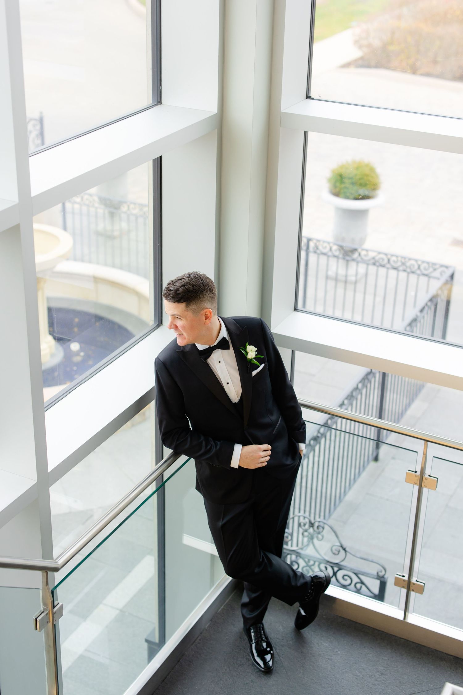 A groom in a classic black tuxedo poses by large windows in a modern building.