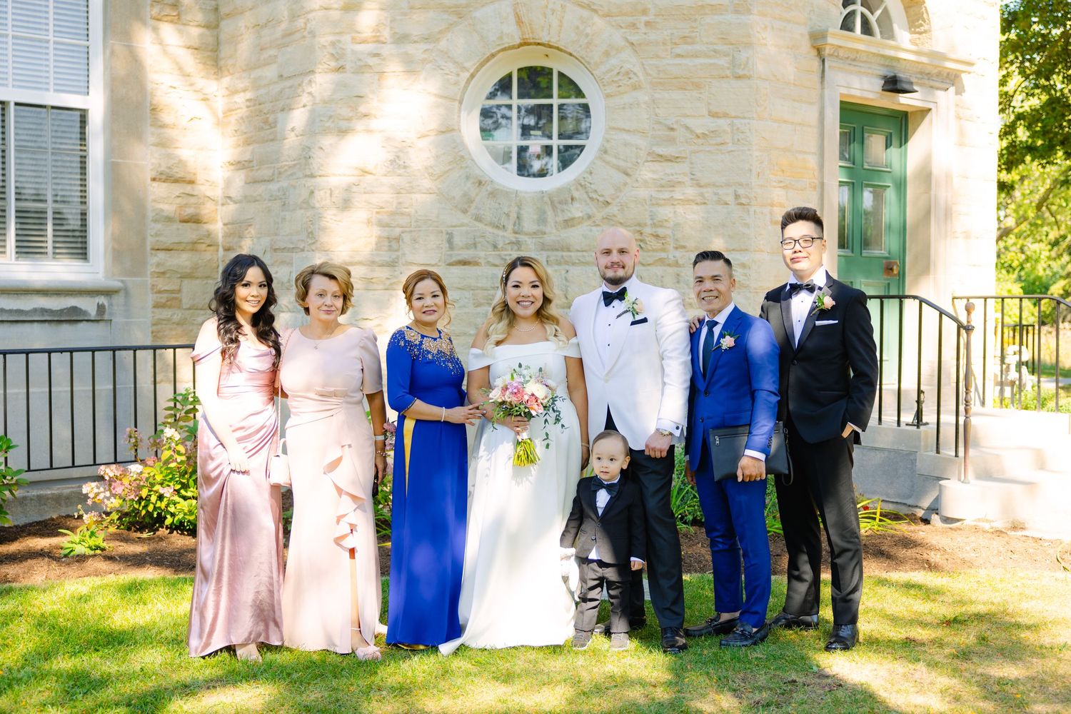 Large wedding party with diverse formal attire poses outside stone church with round window.