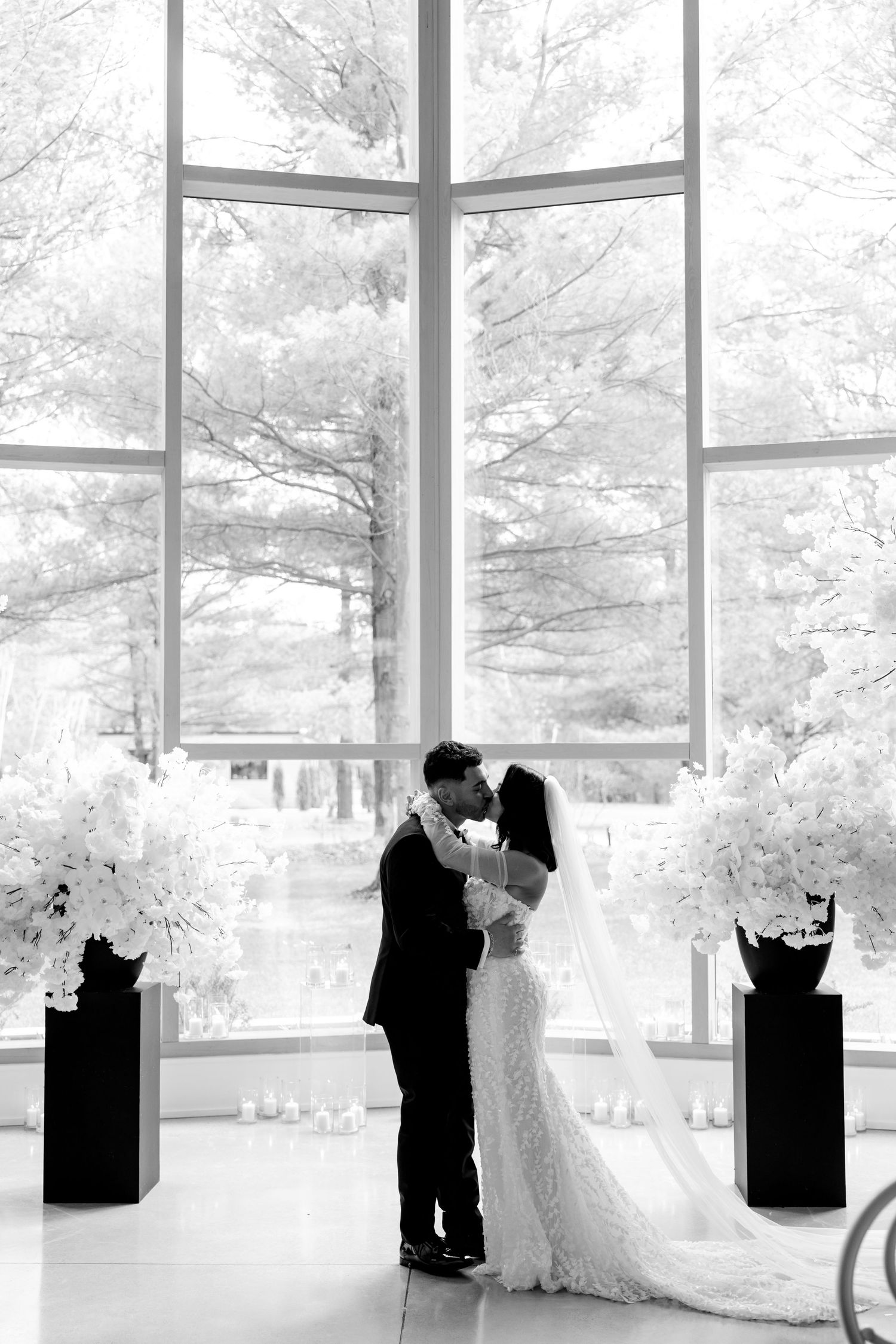 Black and white photo of romantic moment in front of large windows with white floral arrangements.