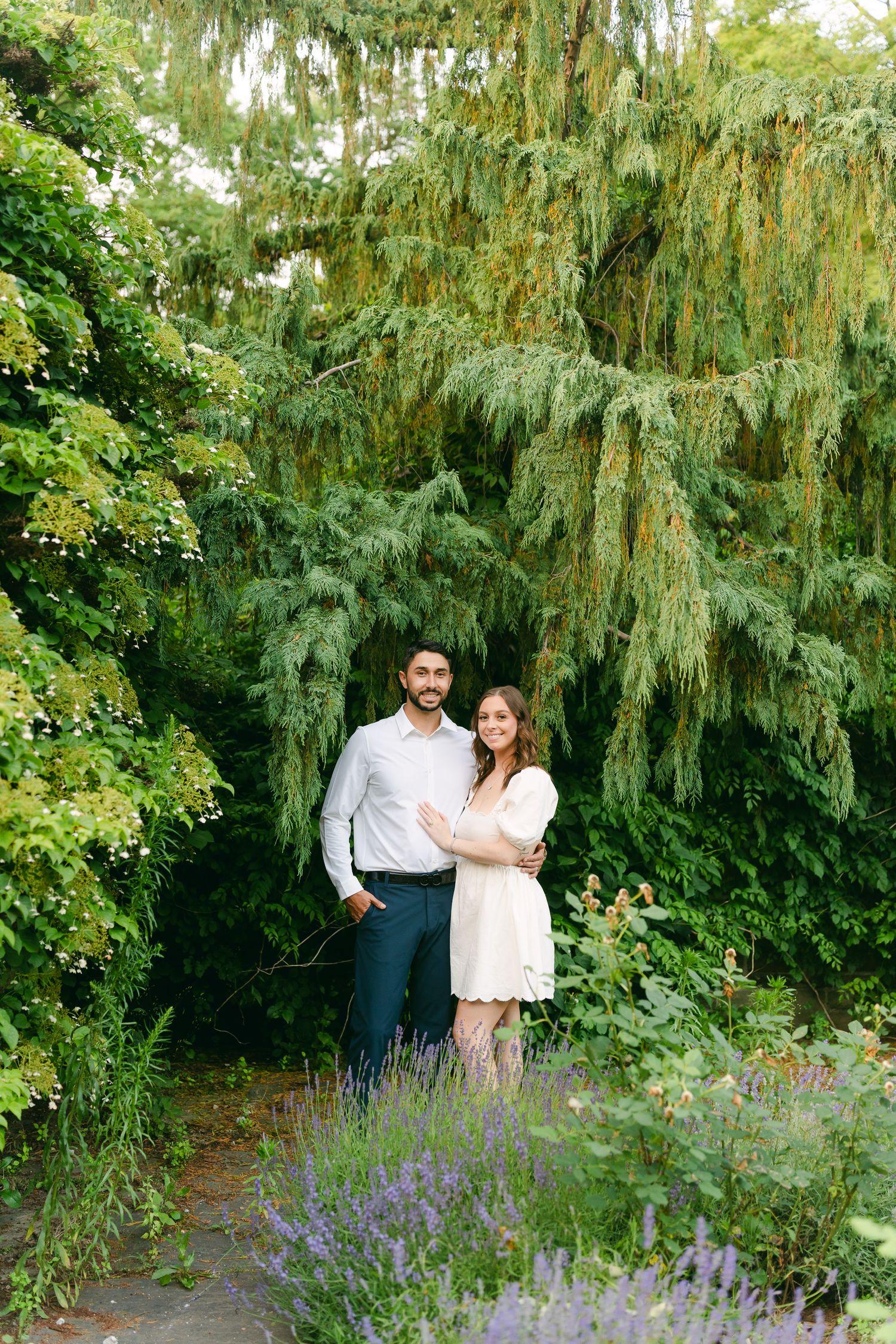 Two people embrace in a romantic garden setting with a weeping willow tree and purple flowers in the foreground.