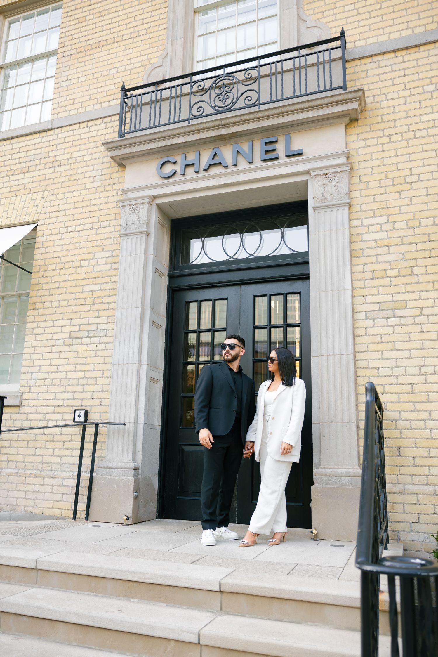 A fashionable couple poses in front of an elegant Chanel storefront entrance.