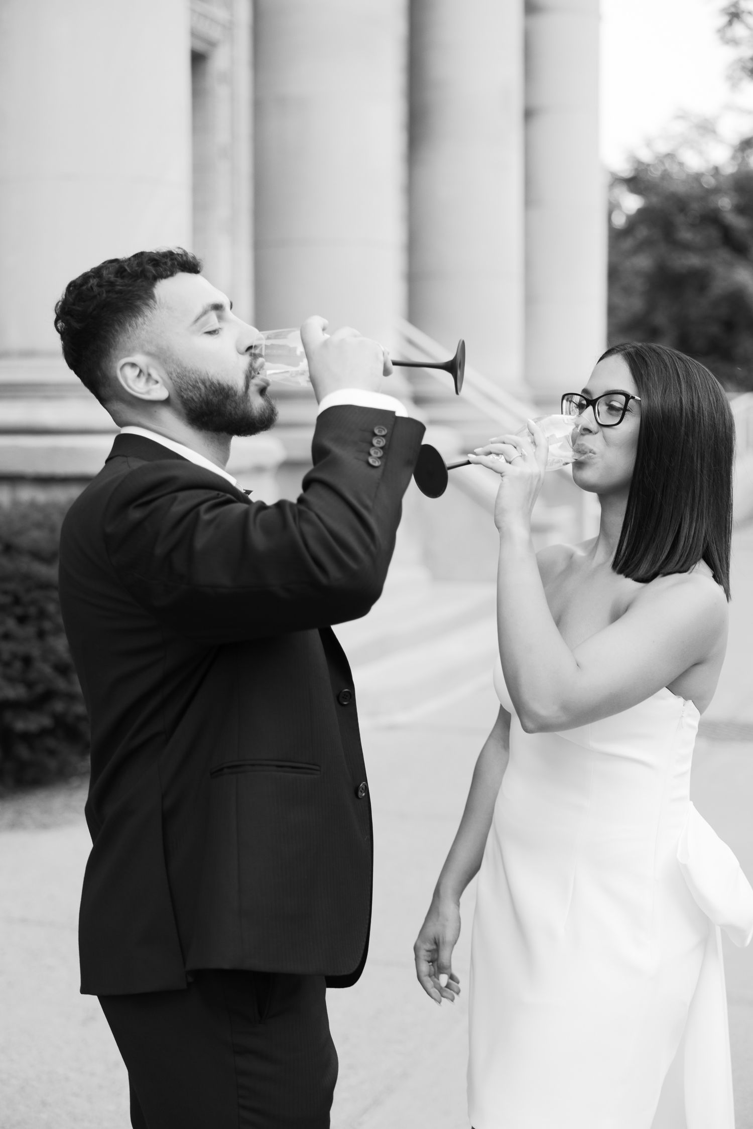 Black and white photo of a couple playfully drinking champagne in front of stone columns.