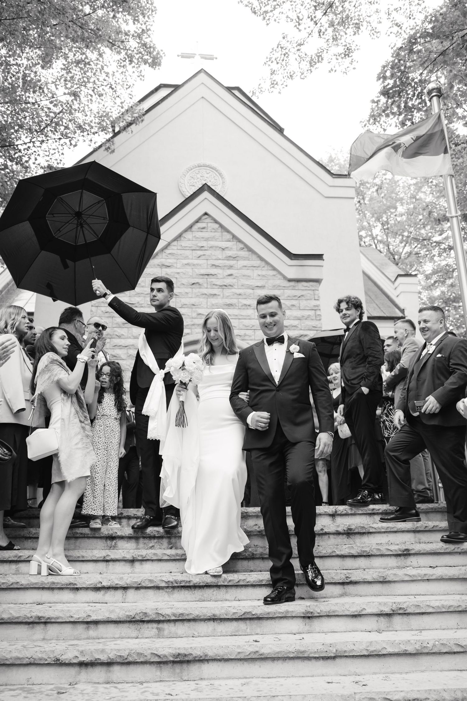 A wedding party descends church steps with black umbrellas on an overcast day.