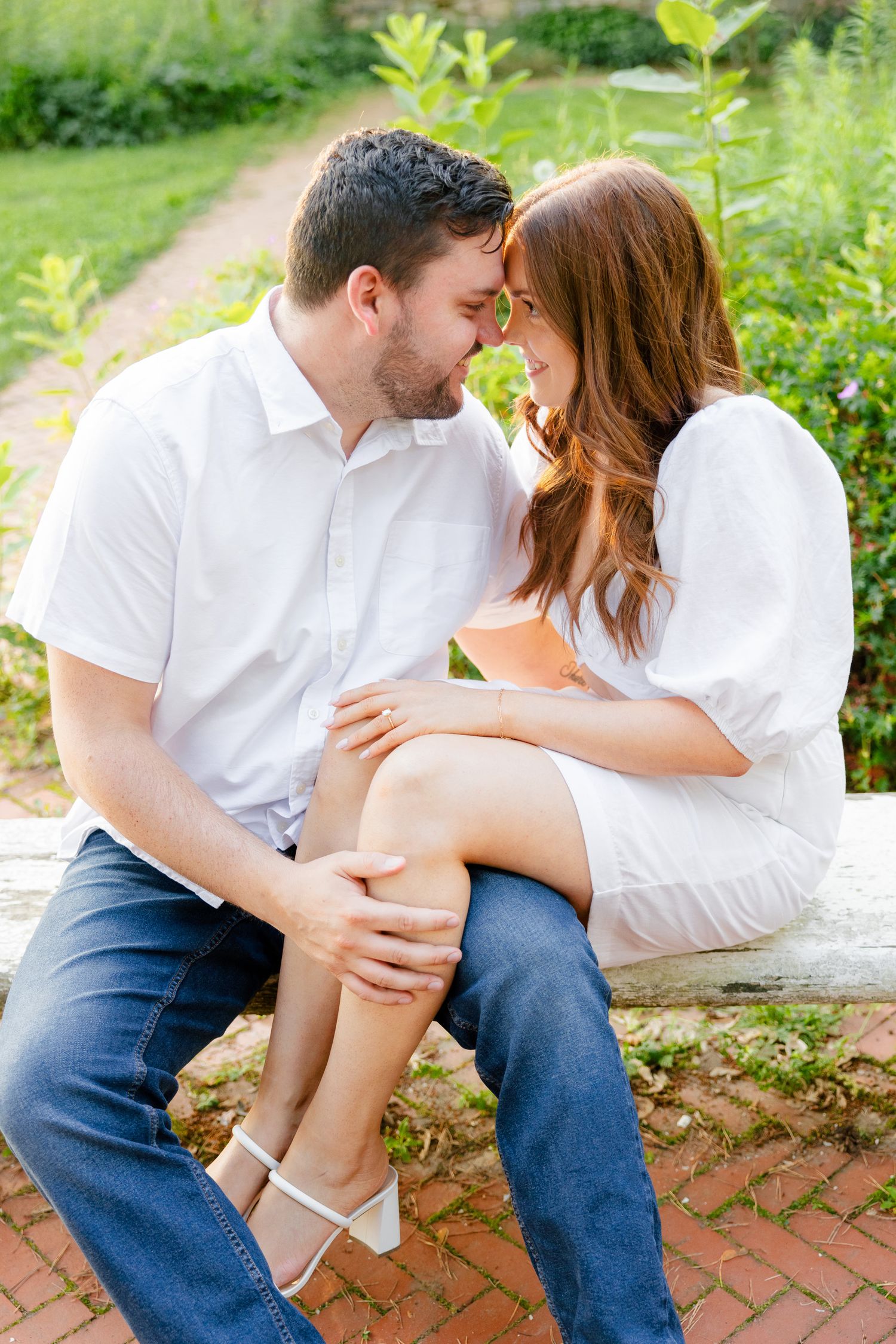 A couple dressed in matching white outfits shares a tender moment in a sunlit garden.