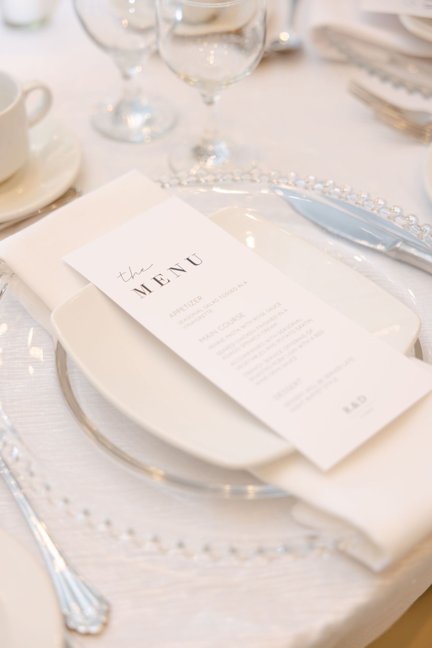 Elegant place setting with crystal charger plate, menu card, and silver cutlery on white tablecloth.