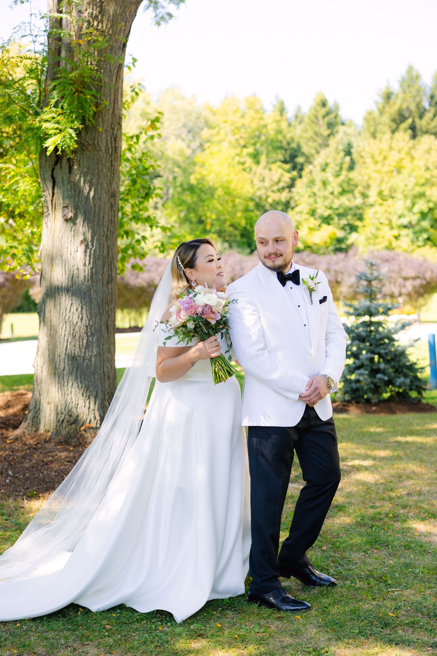 Wedding couple posing outdoors under a tree in a garden with bride holding a pink and white bouquet.