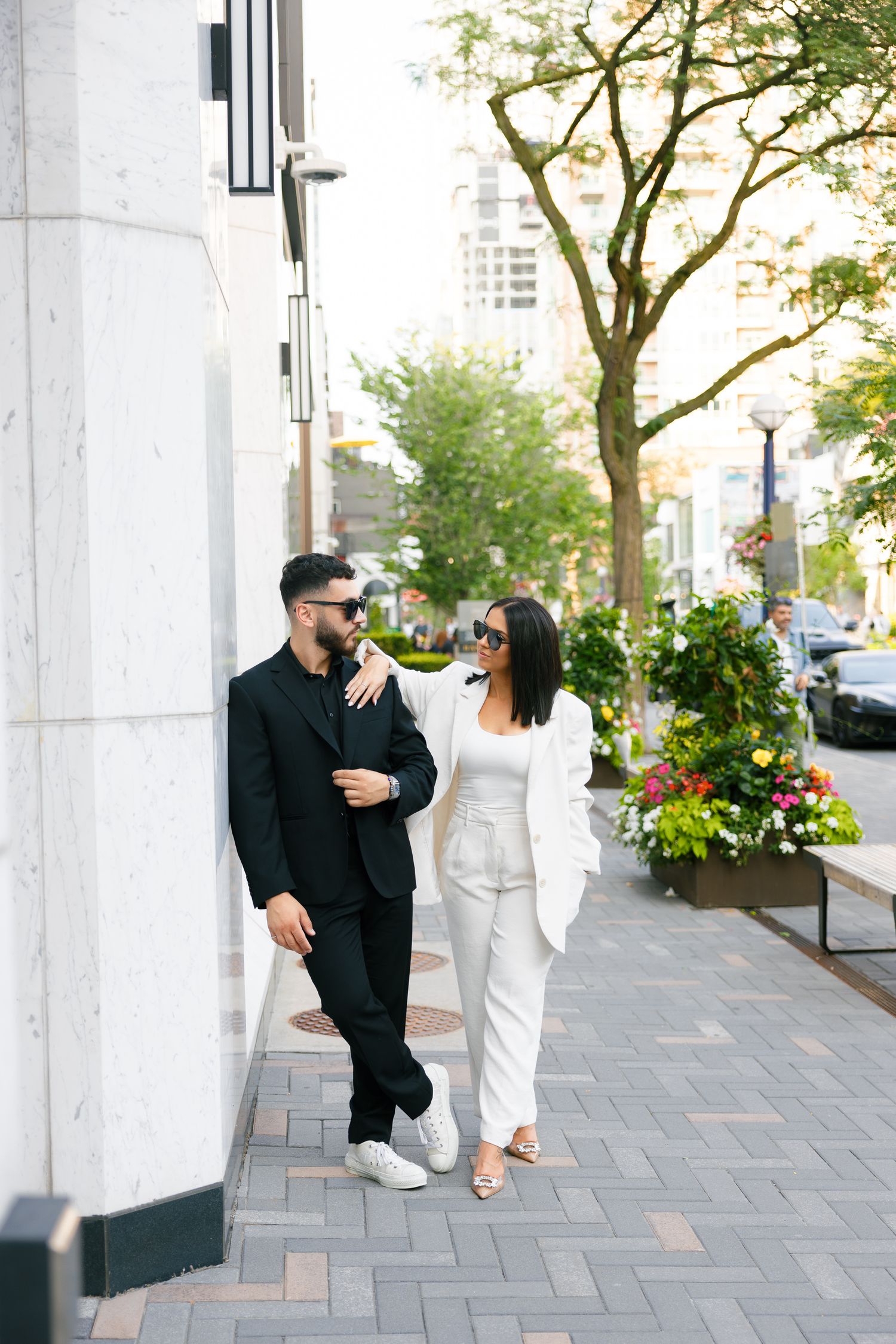 Stylish couple in contrasting black and white outfits stroll together on an urban sidewalk lined with trees.