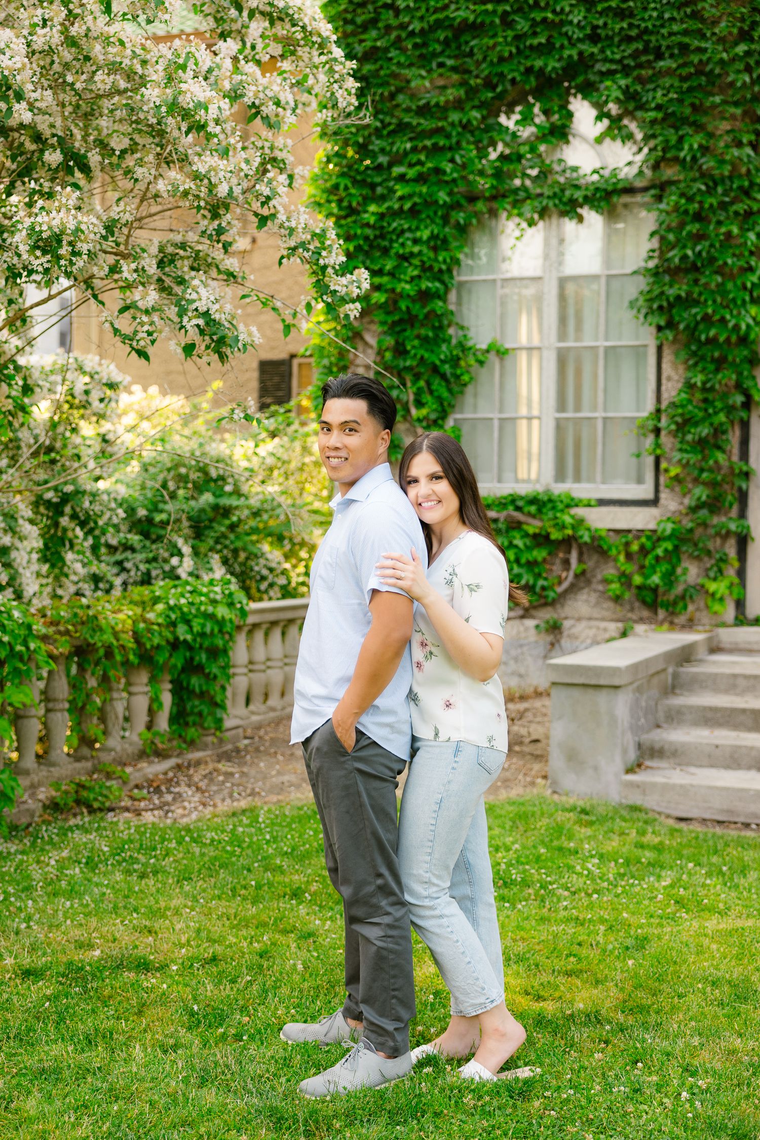 Couple poses back-to-back on lawn with ivy-covered building and blooming trees in background.