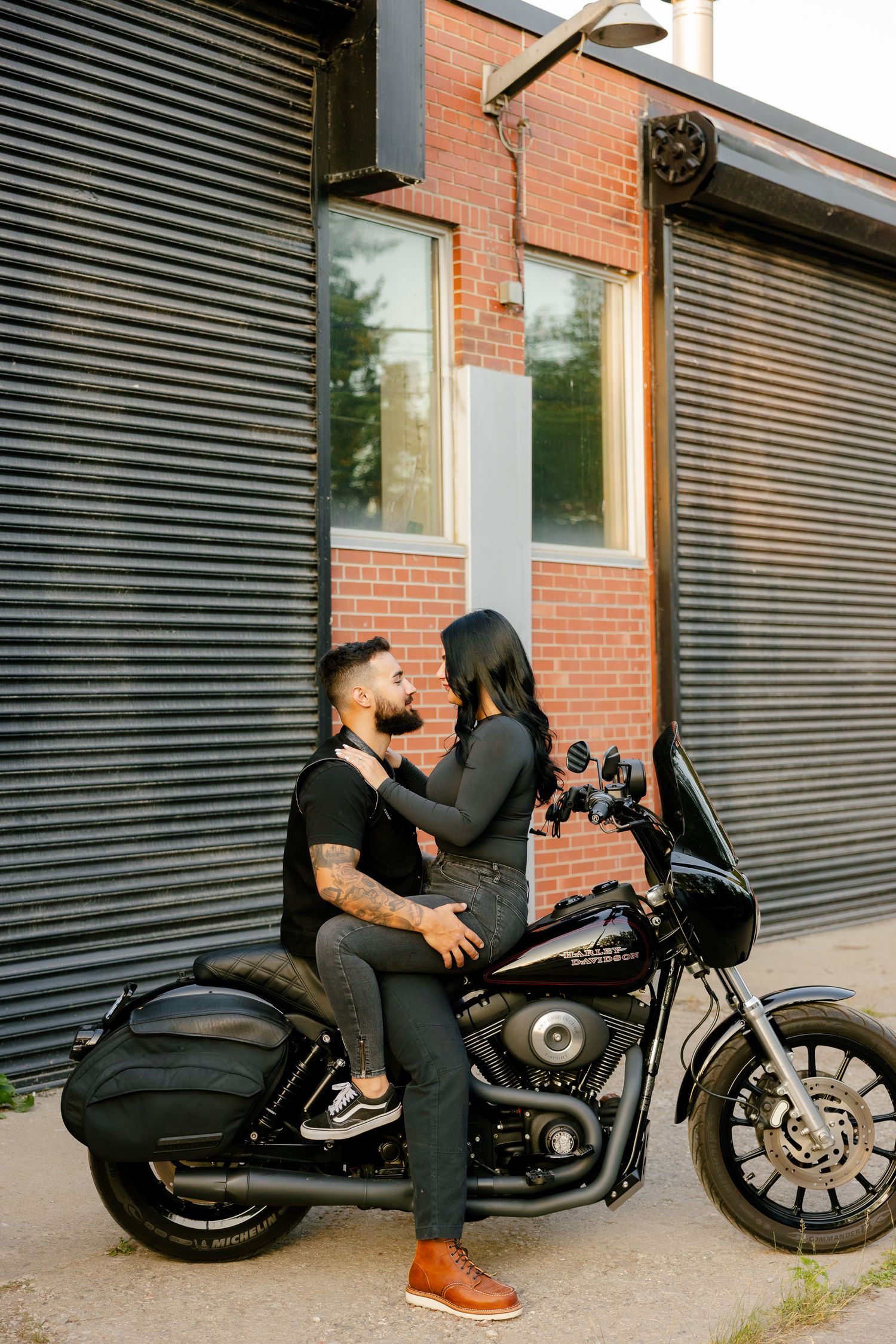 A romantic couple shares an intimate moment on their motorcycle against brick and metal walls.