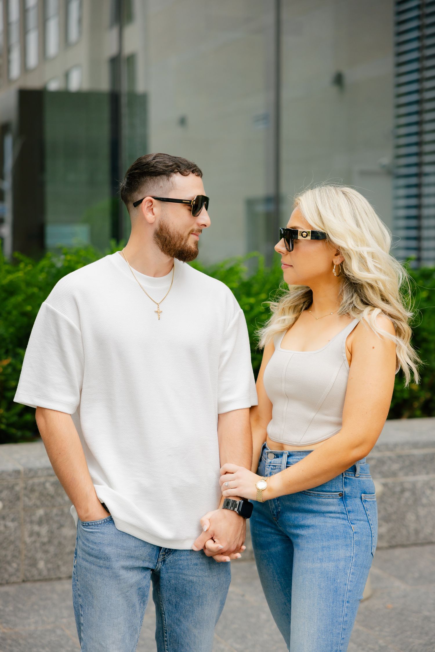 A stylish couple in casual wear and sunglasses pose together outside a modern building.