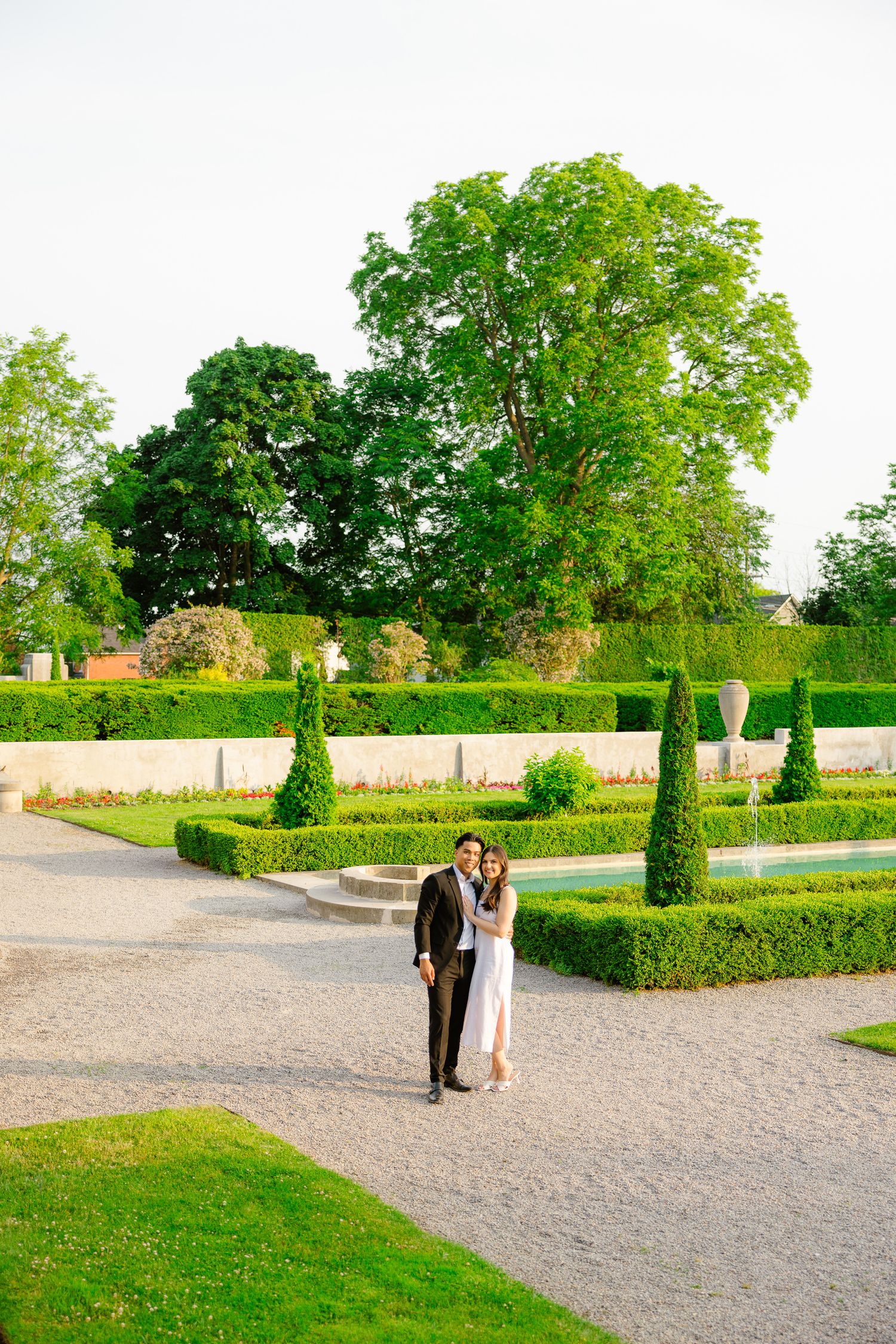 Two people stand together on a garden path surrounded by sculptured hedges and cypress trees.