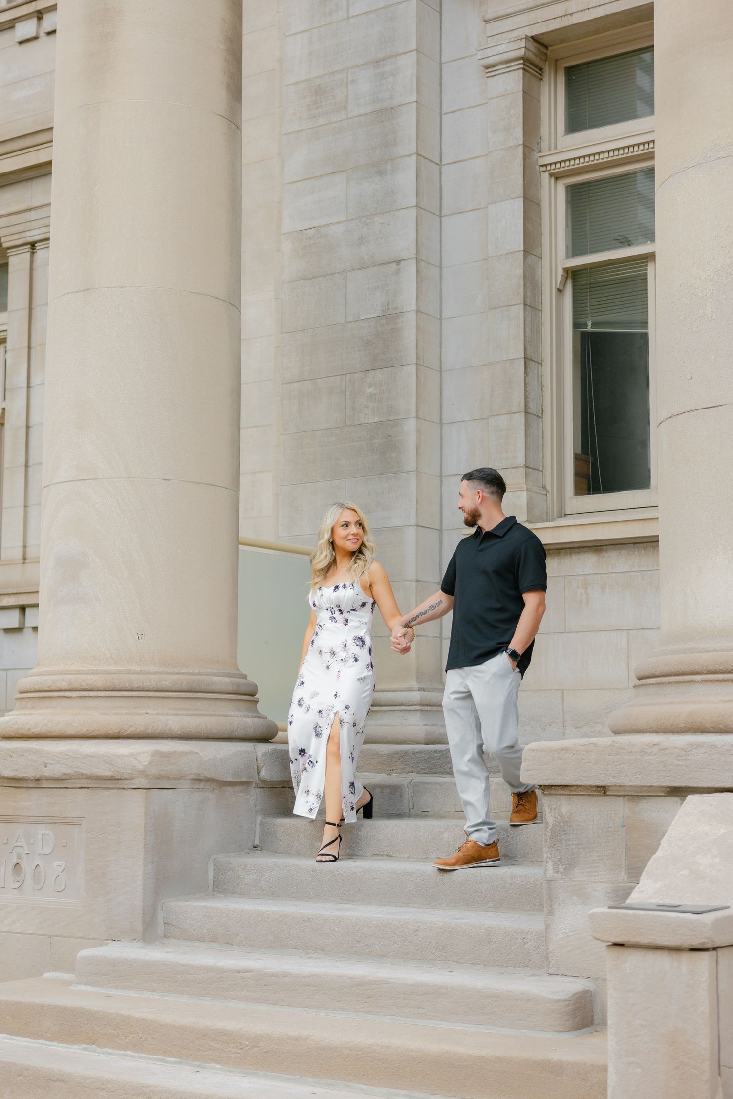 Two people walk down stone steps of a grand building with tall columns and windows.