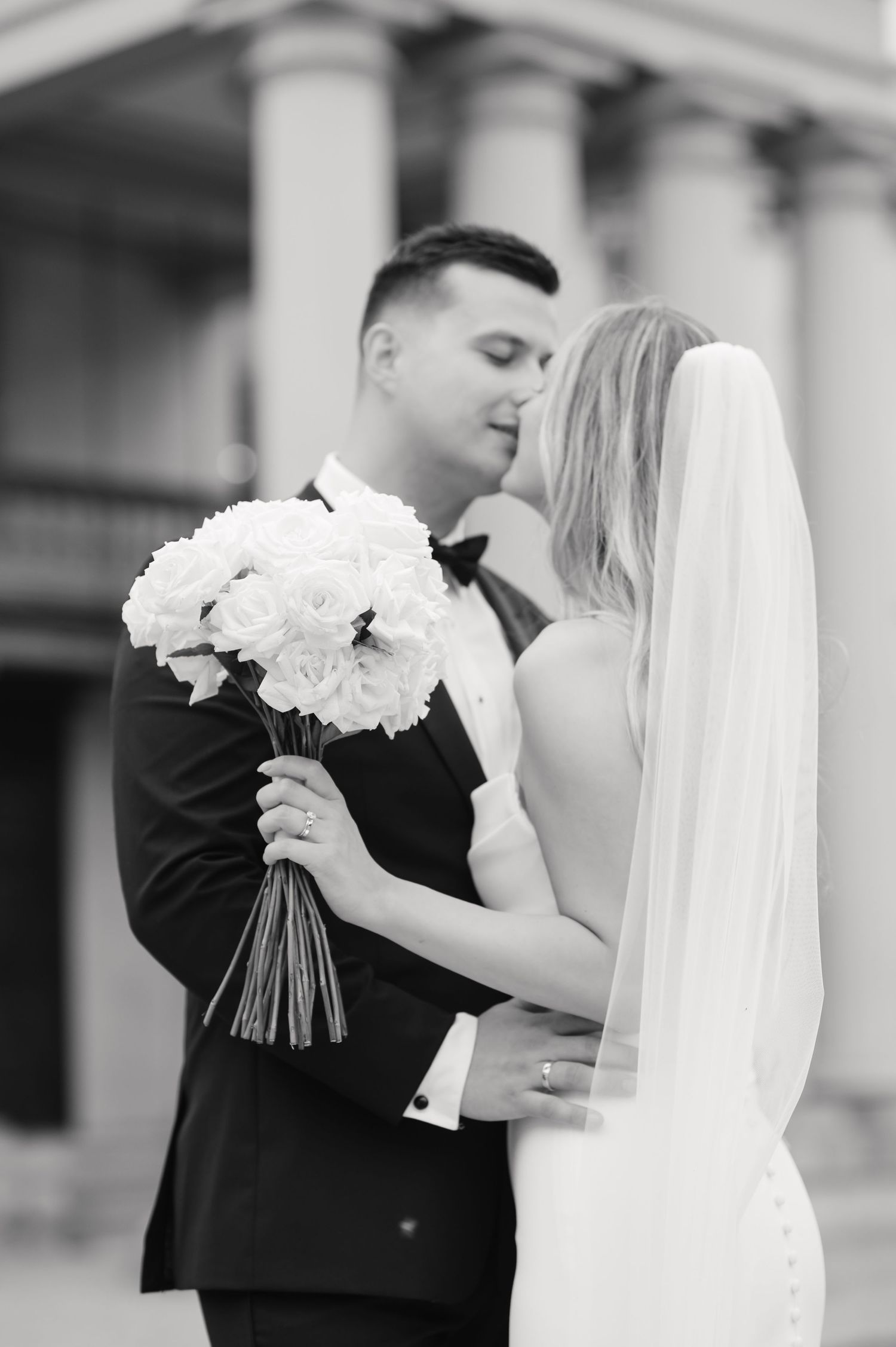 Black and white romantic moment with bride holding white flower bouquet against columned backdrop.