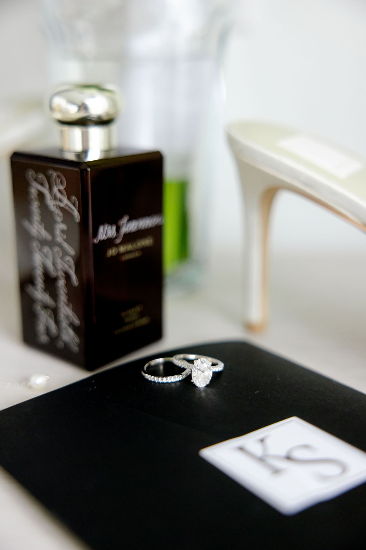 Engagement rings displayed with perfume bottle and white heel on dark surface.
