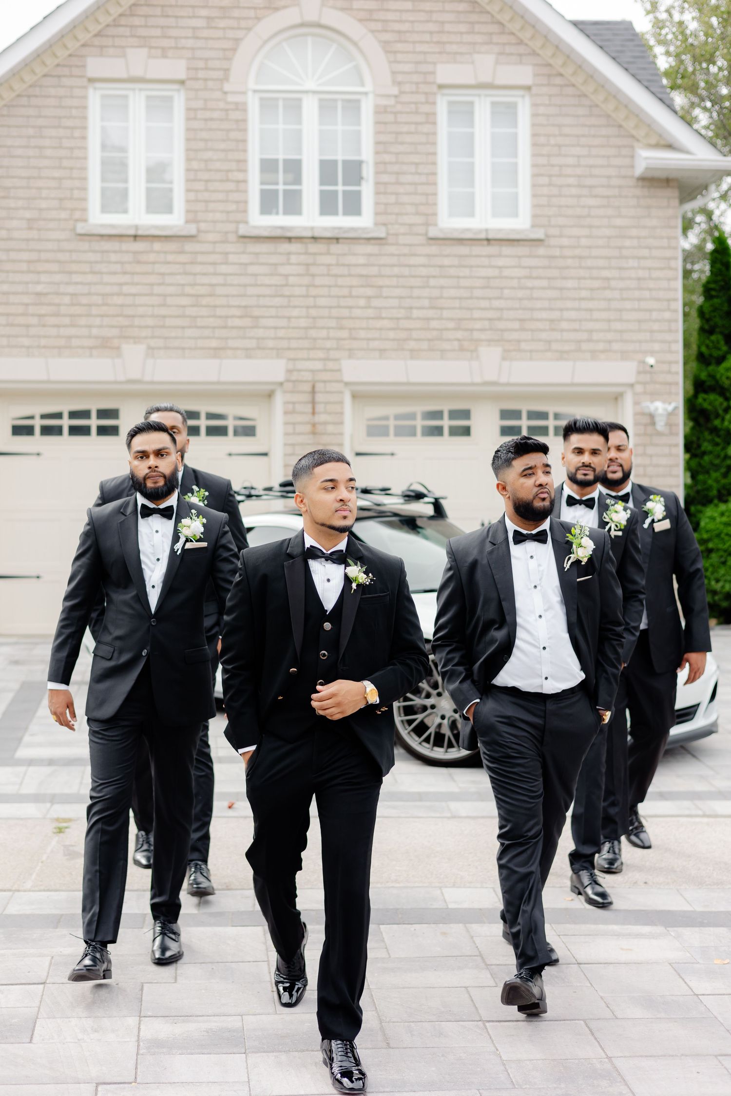 Groomsmen walking in coordinated black tuxedos with white boutonnières outside suburban home.