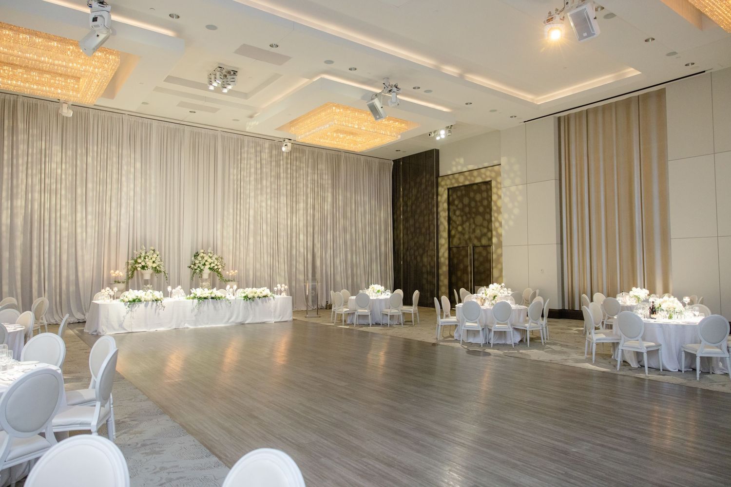 Modern reception venue with white draped walls, elegant chandeliers and round dining tables.