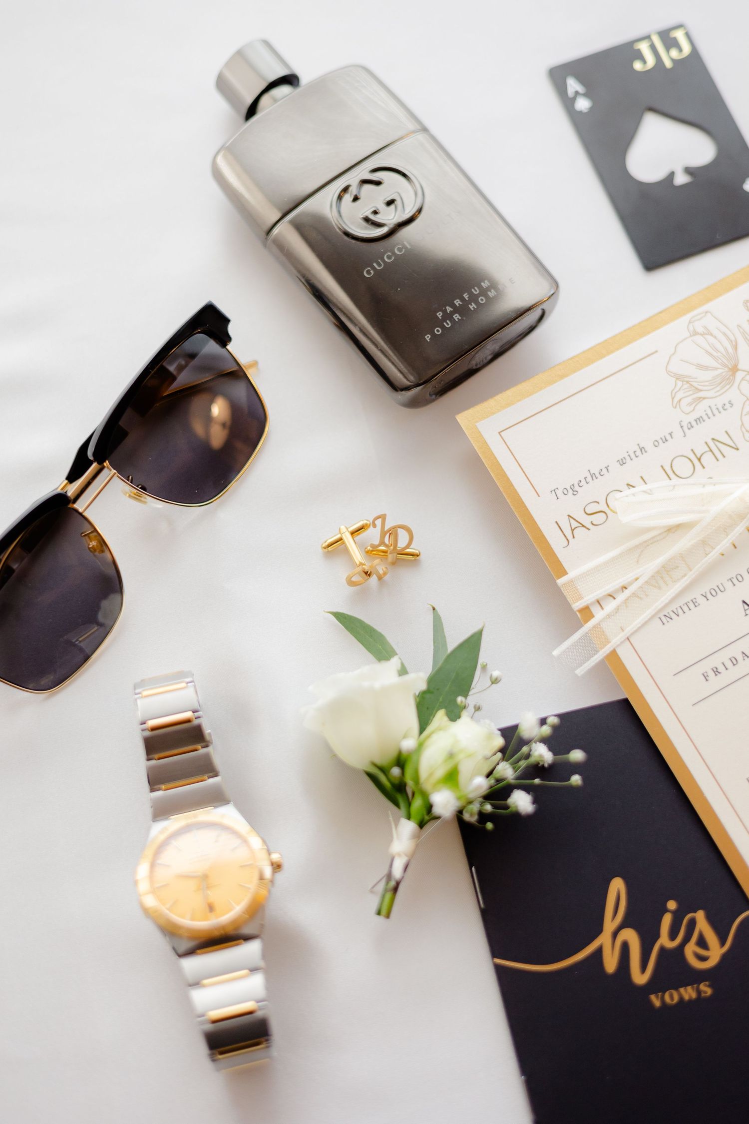 Luxury wedding details including sunglasses, gold watch, boutonniere and invitation on white surface.