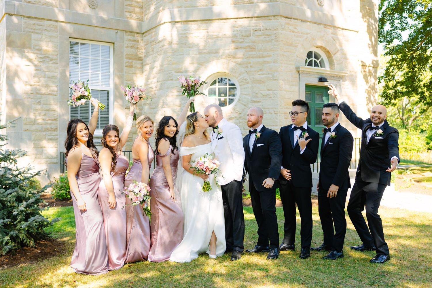 Wedding party with bridesmaids in pink dresses and groomsmen in black suits celebrating outdoors.