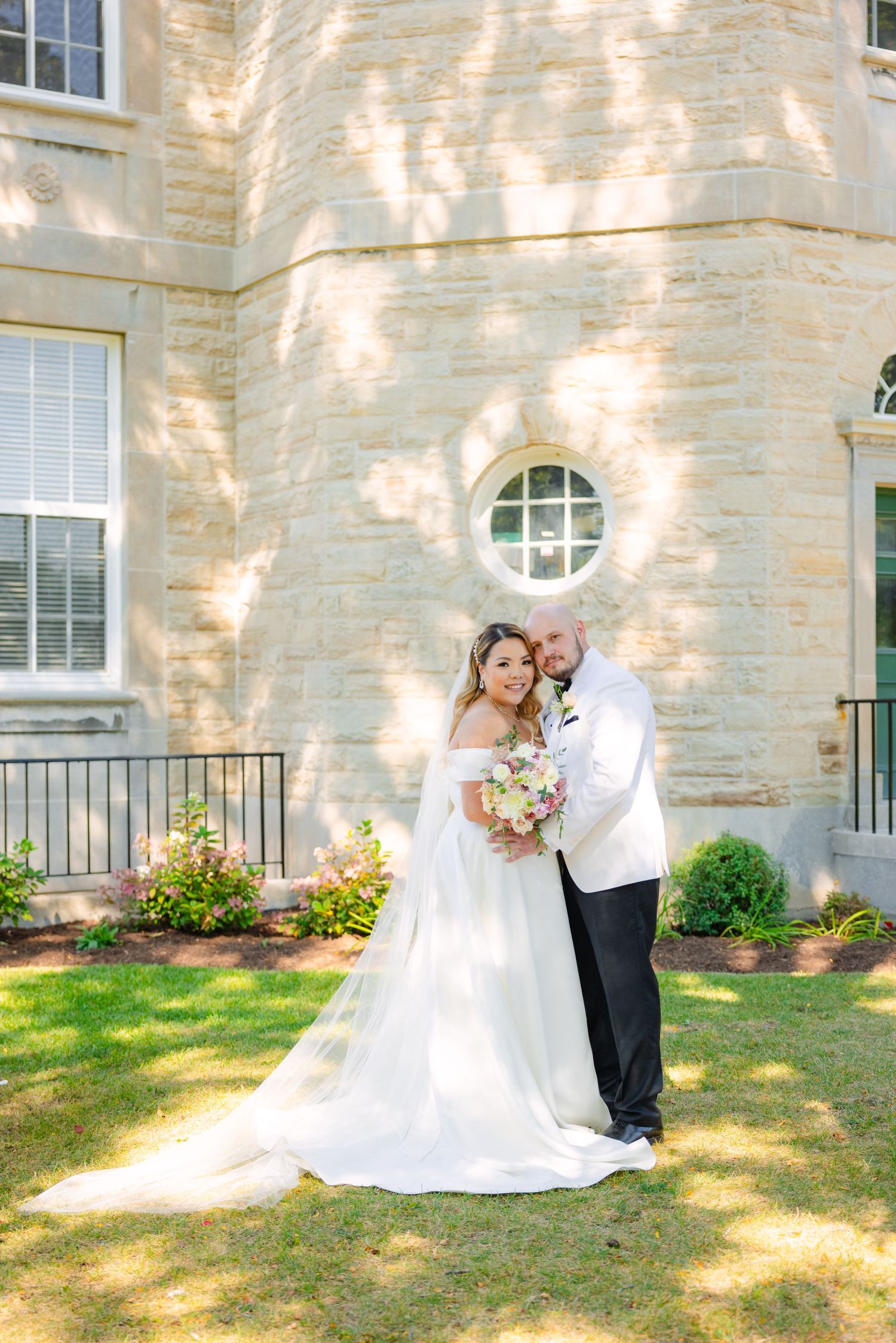 Wedding couple poses elegantly against stone wall with round window on sunny afternoon.