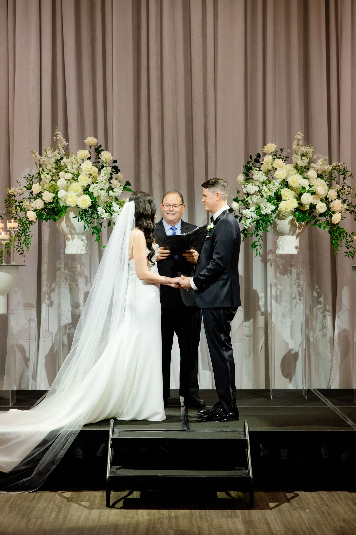 Wedding ceremony on elevated platform with lush white floral arrangements against taupe drapery.