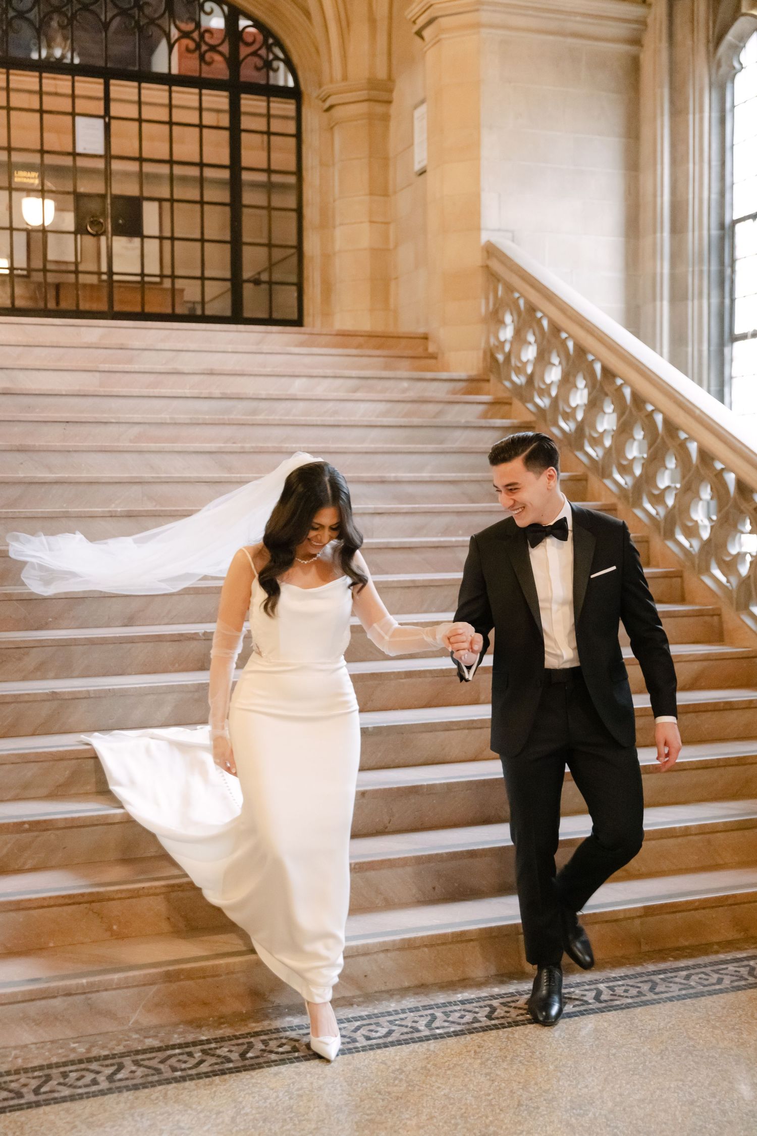 A joyful couple walks down marble stairs in elegant wedding attire beneath vaulted gothic architecture.
