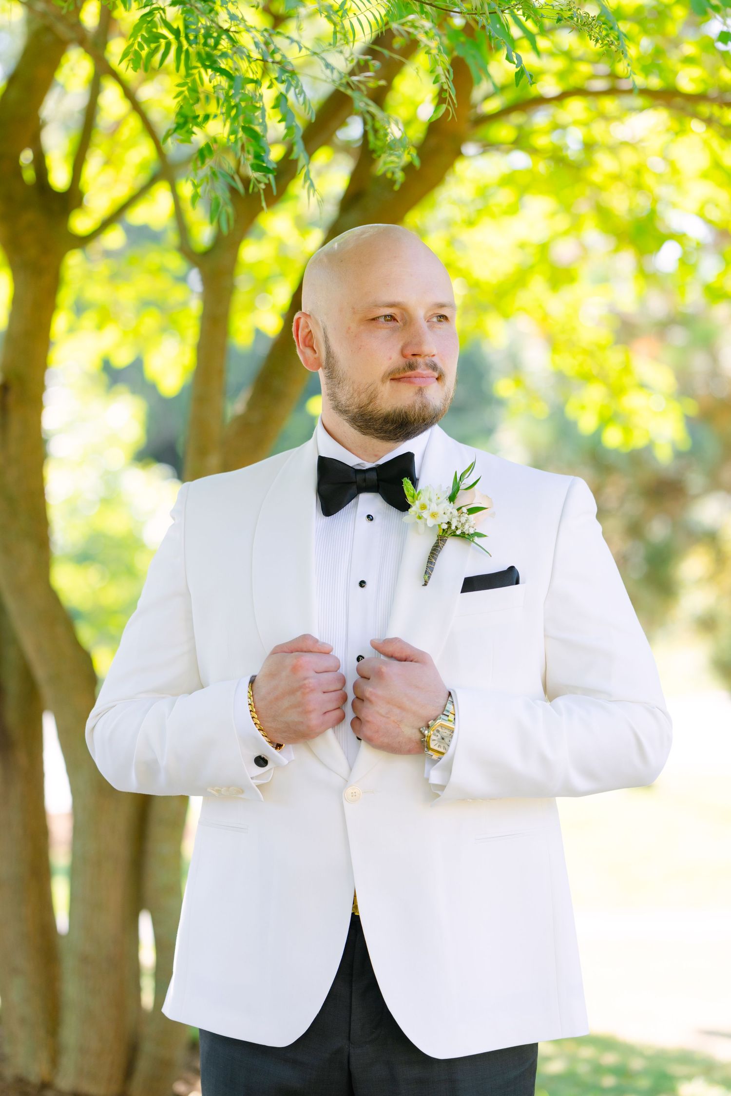 Groom in white tuxedo jacket and black bow tie poses confidently in garden setting.