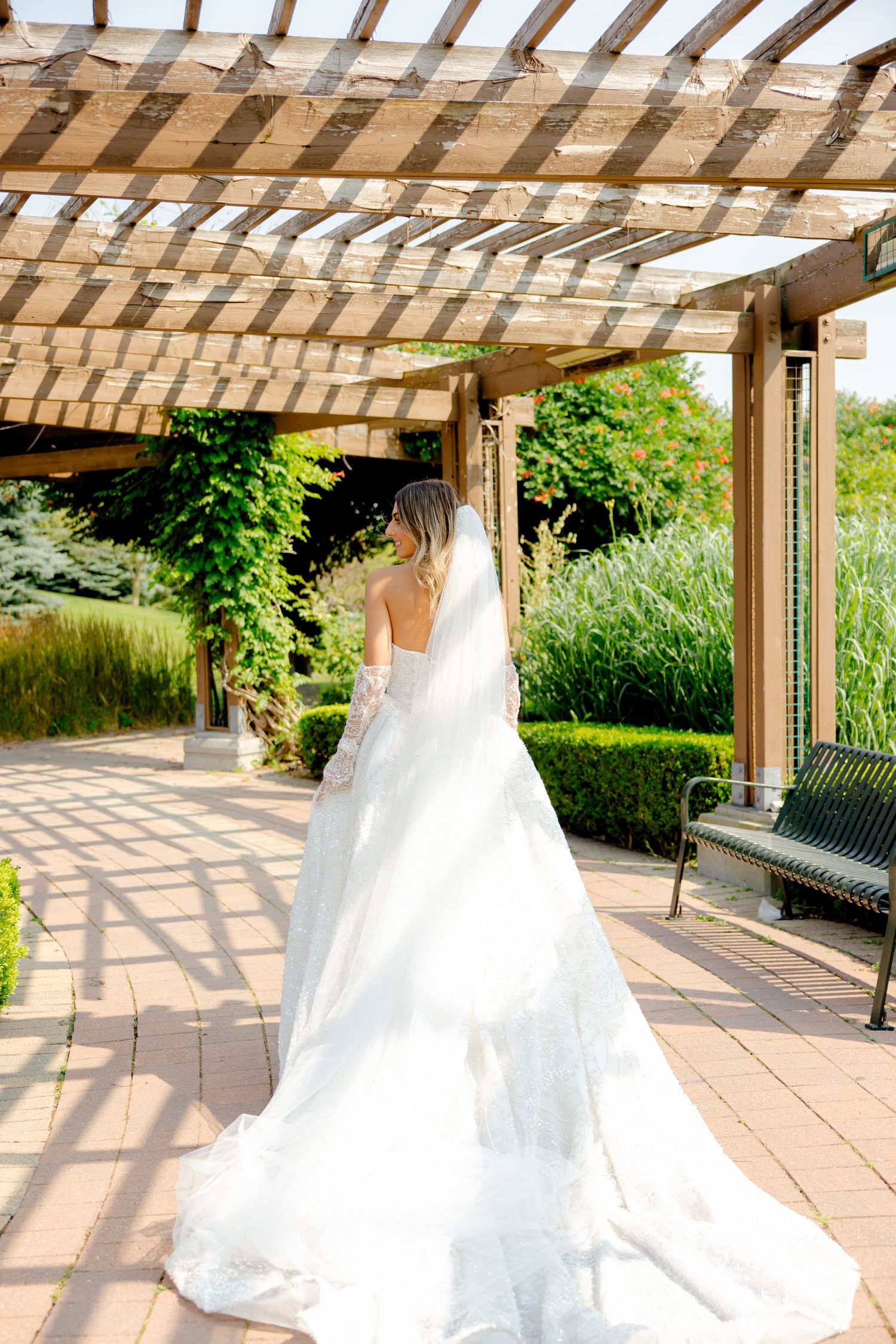 Bride in white gown stands under wooden pergola casting geometric shadows.